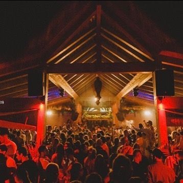 Gran multitud bailando bajo una estructura de madera por la noche; iluminación roja y azul.