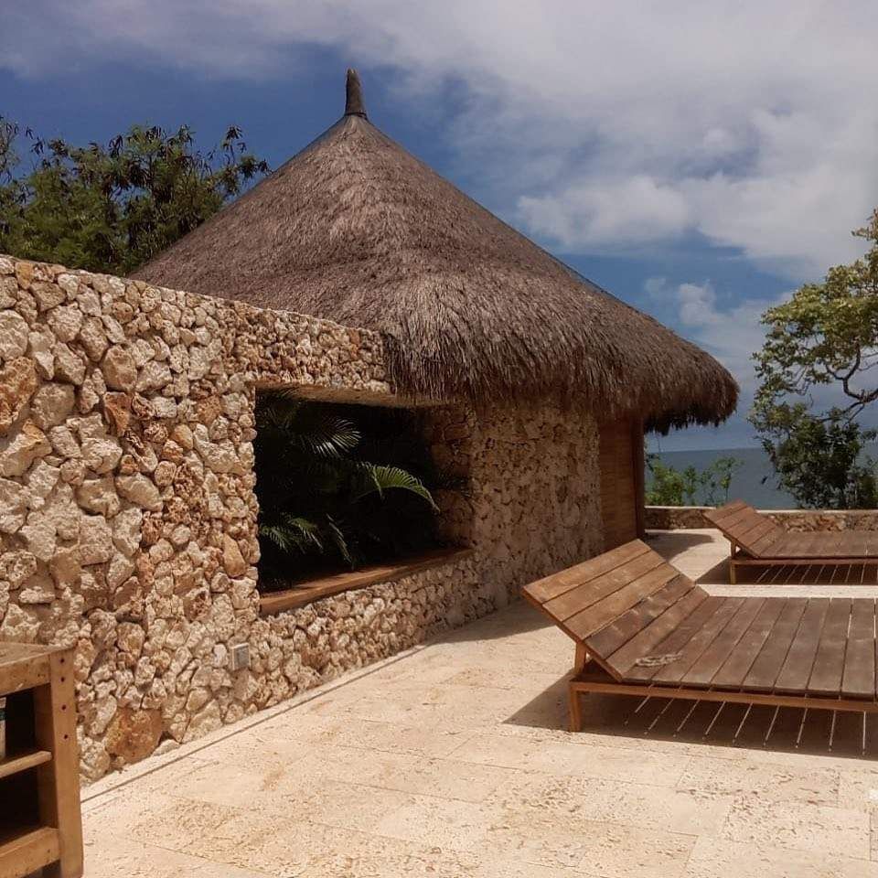 Edificio de piedra con techo de paja, una ventana y tumbonas de madera en un patio junto al agua.