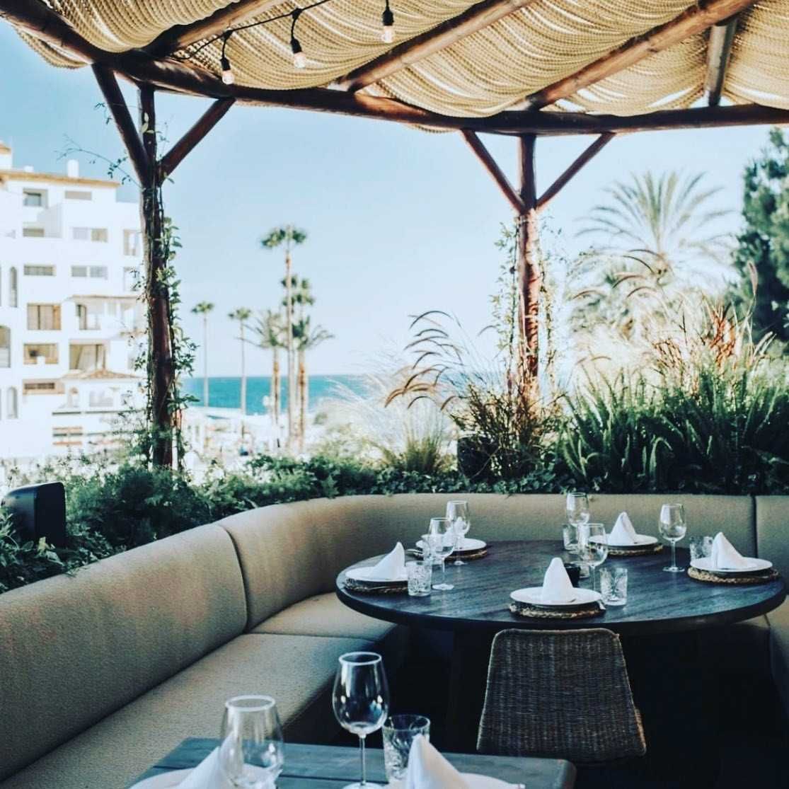 Comedor al aire libre con vista al mar; mesa redonda dispuesta para invitados, cubierta por techo de madera y paja.