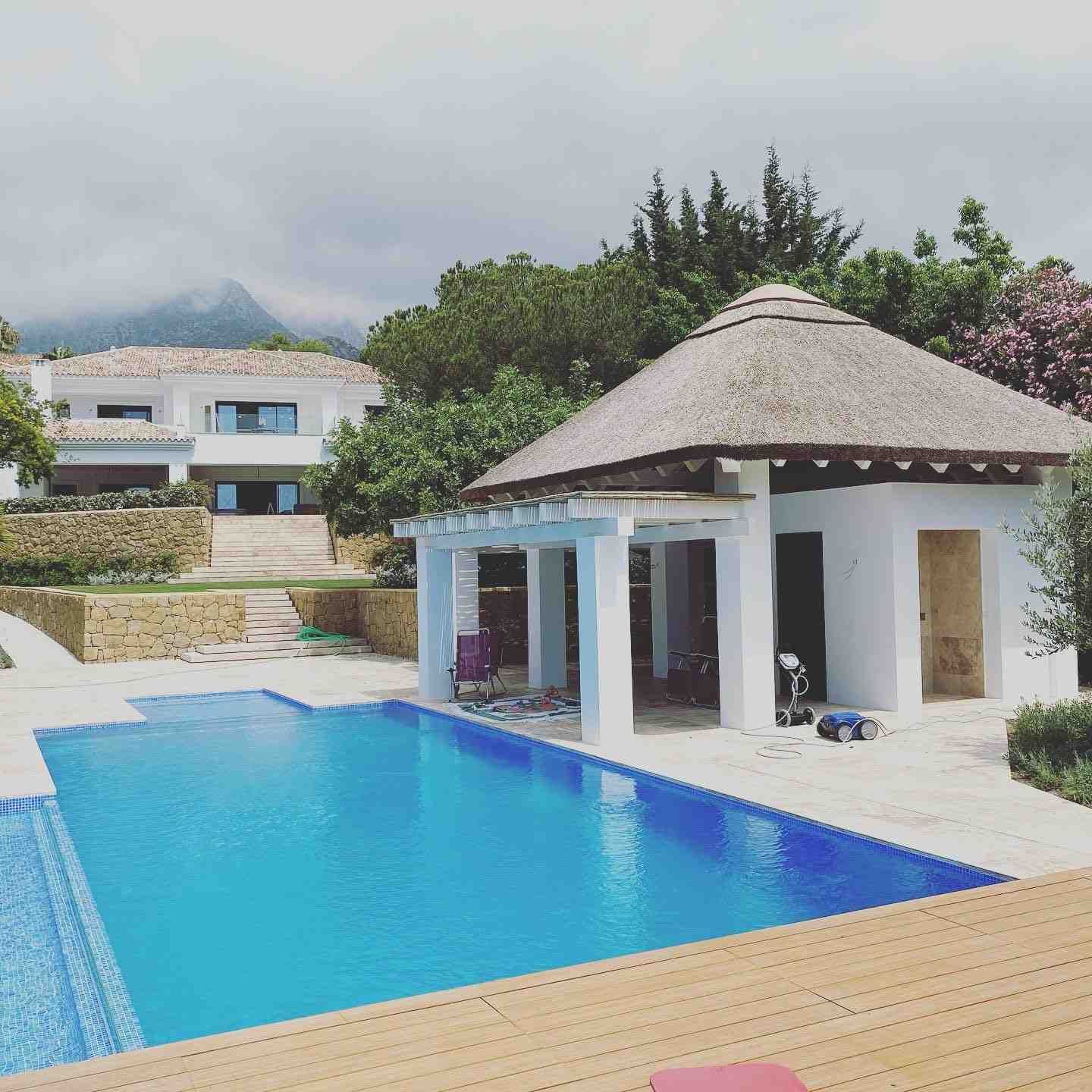 Piscina y cabaña con techo de paja frente a un edificio blanco en un día soleado.