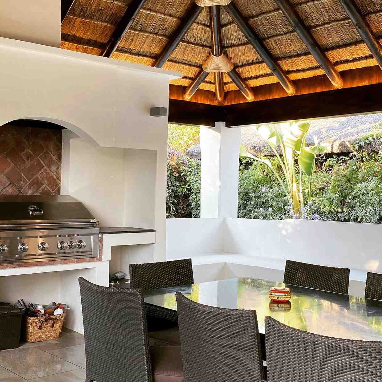 Comedor al aire libre con parrilla y techo de paja, asientos alrededor de una mesa grande y exuberante vegetación.