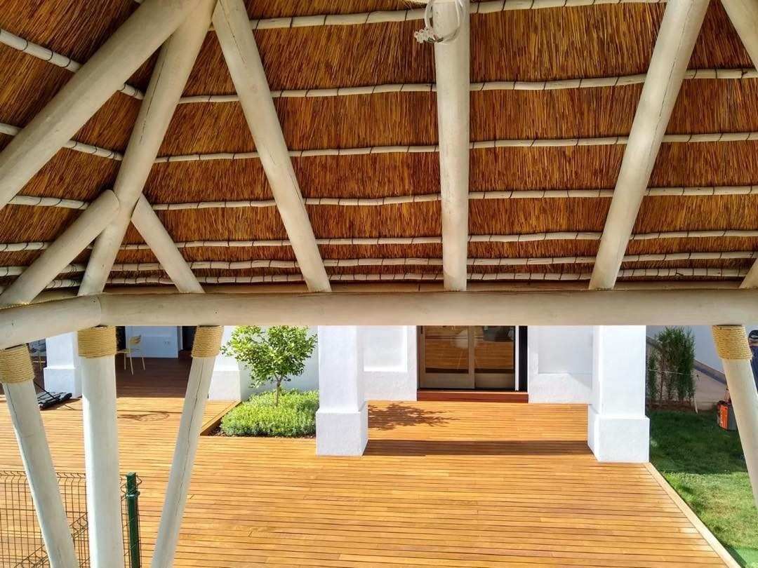 Porche con suelo de madera y techo de paja, sostenido por pilares blancos. Vista hacia un edificio blanco y vegetación.