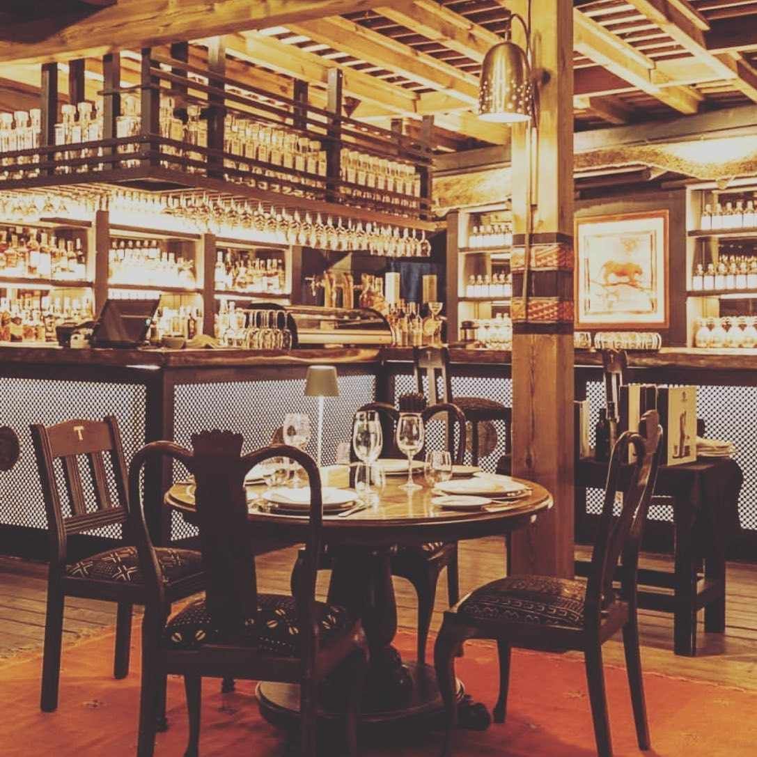 Interior de restaurante con mesas, sillas y barra de madera. La iluminación suave crea un ambiente cálido.