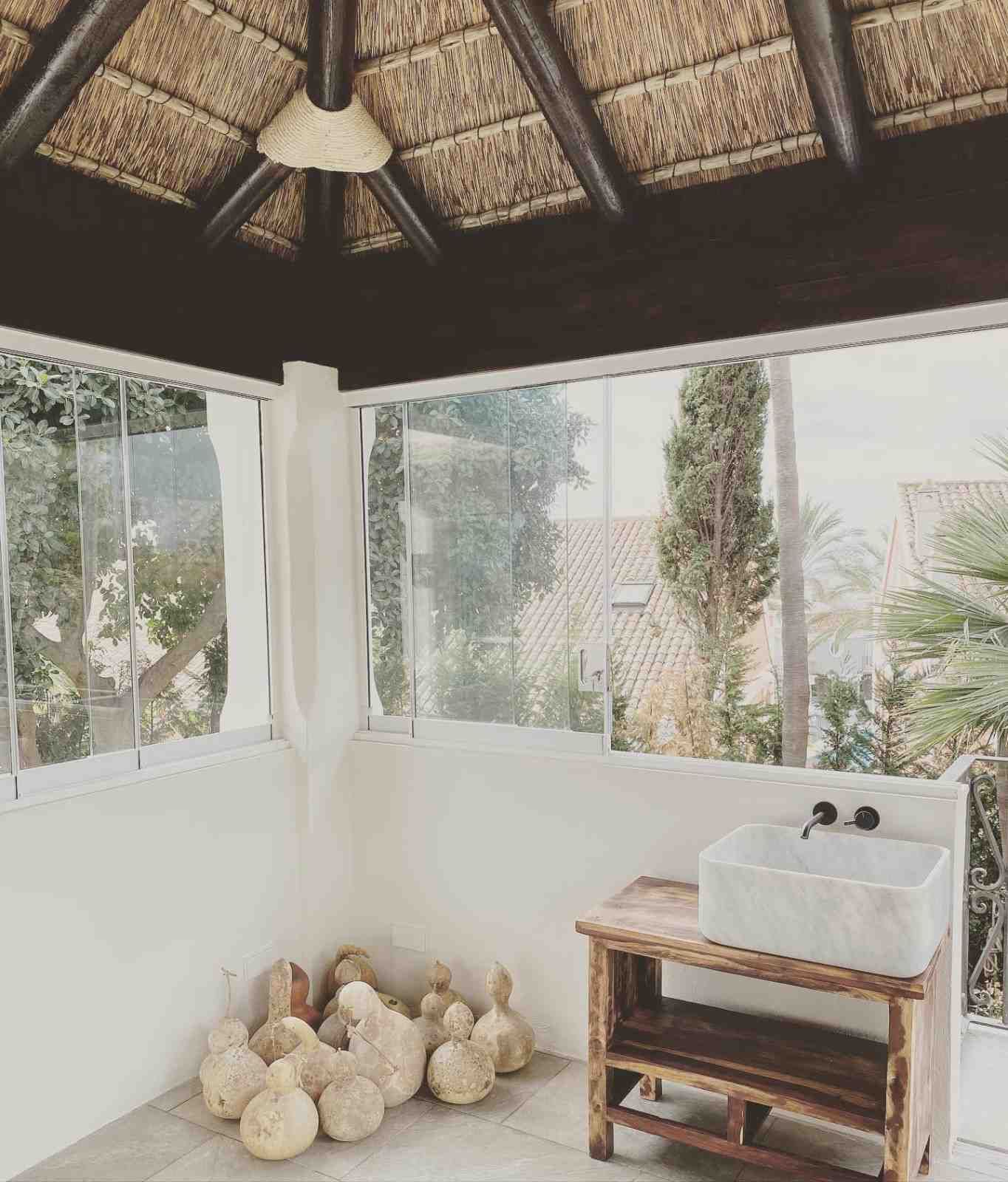 Cenador con techo de paja y ventanas de cristal, lavabo rústico y objetos decorativos de piedra.