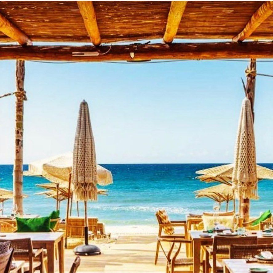 Restaurante frente al mar con vista al mar, mesas, sombrillas y toldo de madera. Aguas azules y cielo soleado.
