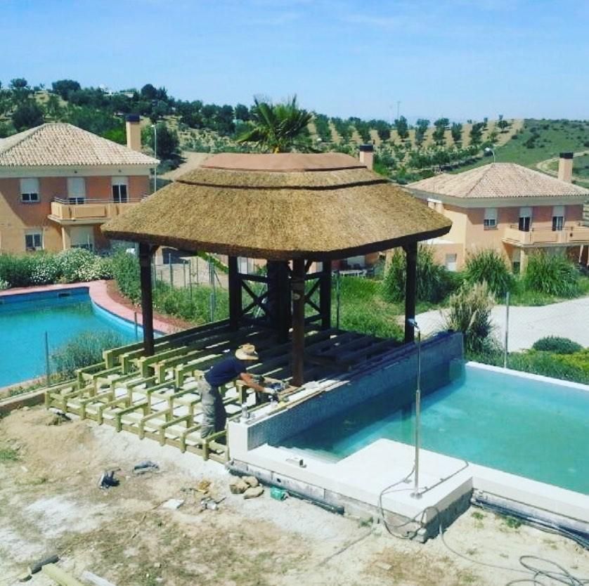 Hombre construyendo una plataforma de madera alrededor de una piscina con un mirador con techo de paja; día soleado.