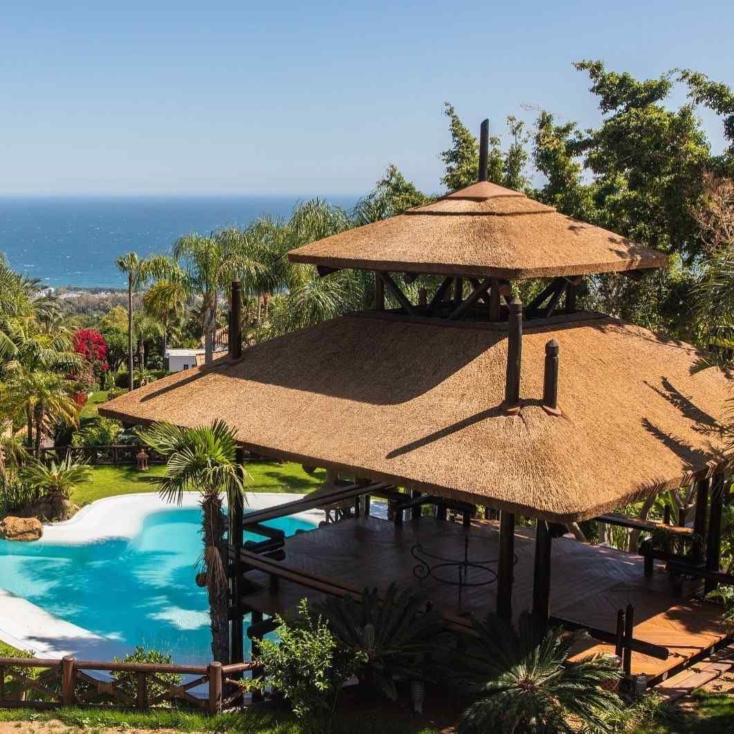 Gazebo con techo de paja con vistas a la piscina y al océano. Está rodeado de exuberante vegetación.