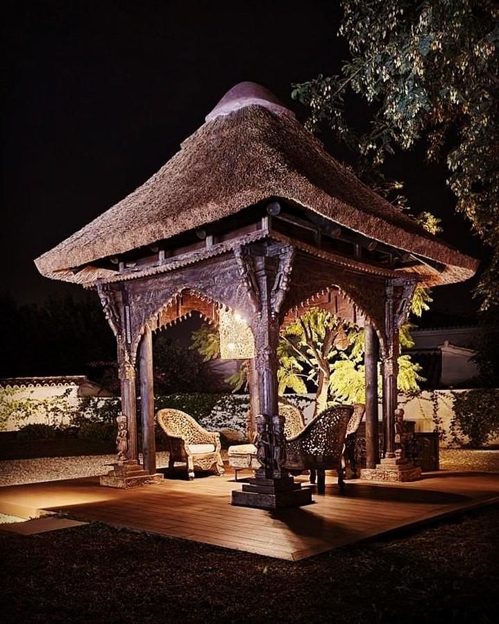 Cenador iluminado con techo de paja, tallas de madera ornamentadas, dos sillas de mimbre en el interior.