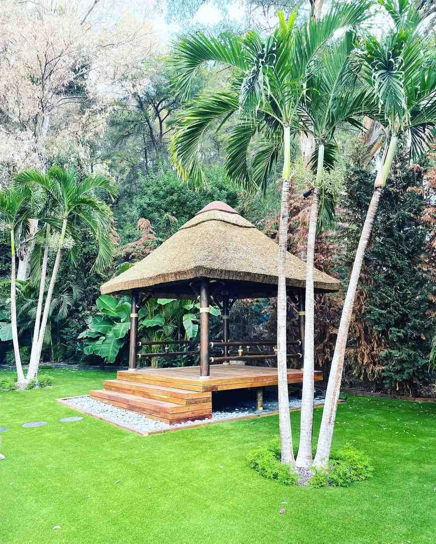 Gazebo con techo de paja en un jardín tropical rodeado de palmeras y vegetación.