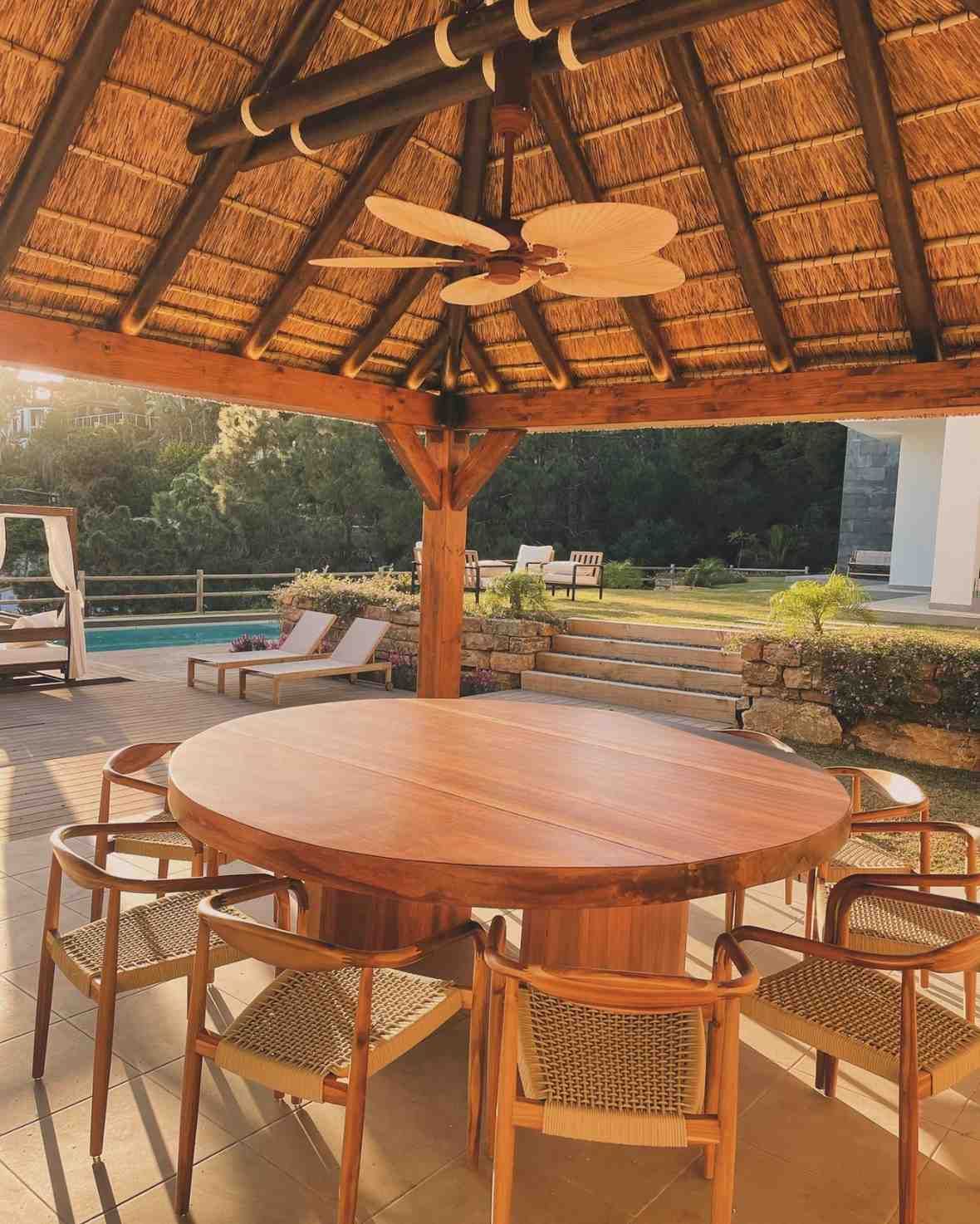 Comedor exterior: gran mesa redonda de madera, sillas, techo de paja, piscina al fondo.