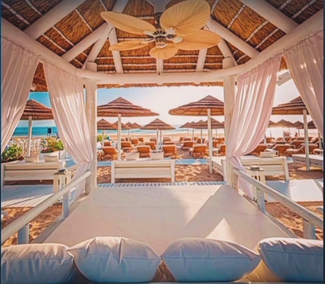 Gazebo en una playa con cortinas blancas, tumbonas y sombrillas de paja.
