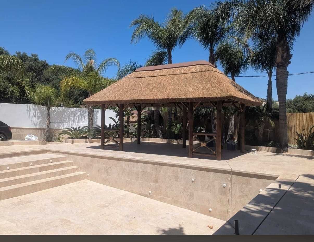 Gazebo con techo de paja cerca de una piscina en construcción, con palmeras y un cielo azul claro.