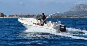 Una persona conduciendo una lancha motora blanca a través del mar, con un pueblo costero y montañas al fondo.