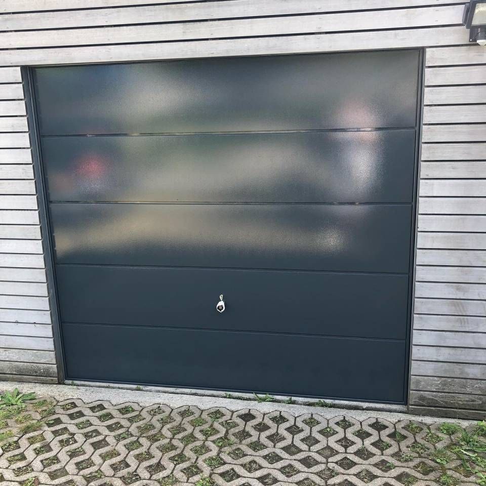 Porta da garage grigio scuro in un edificio con pareti in legno, con maniglia argentata. Vialetto d'accesso in mattoni.