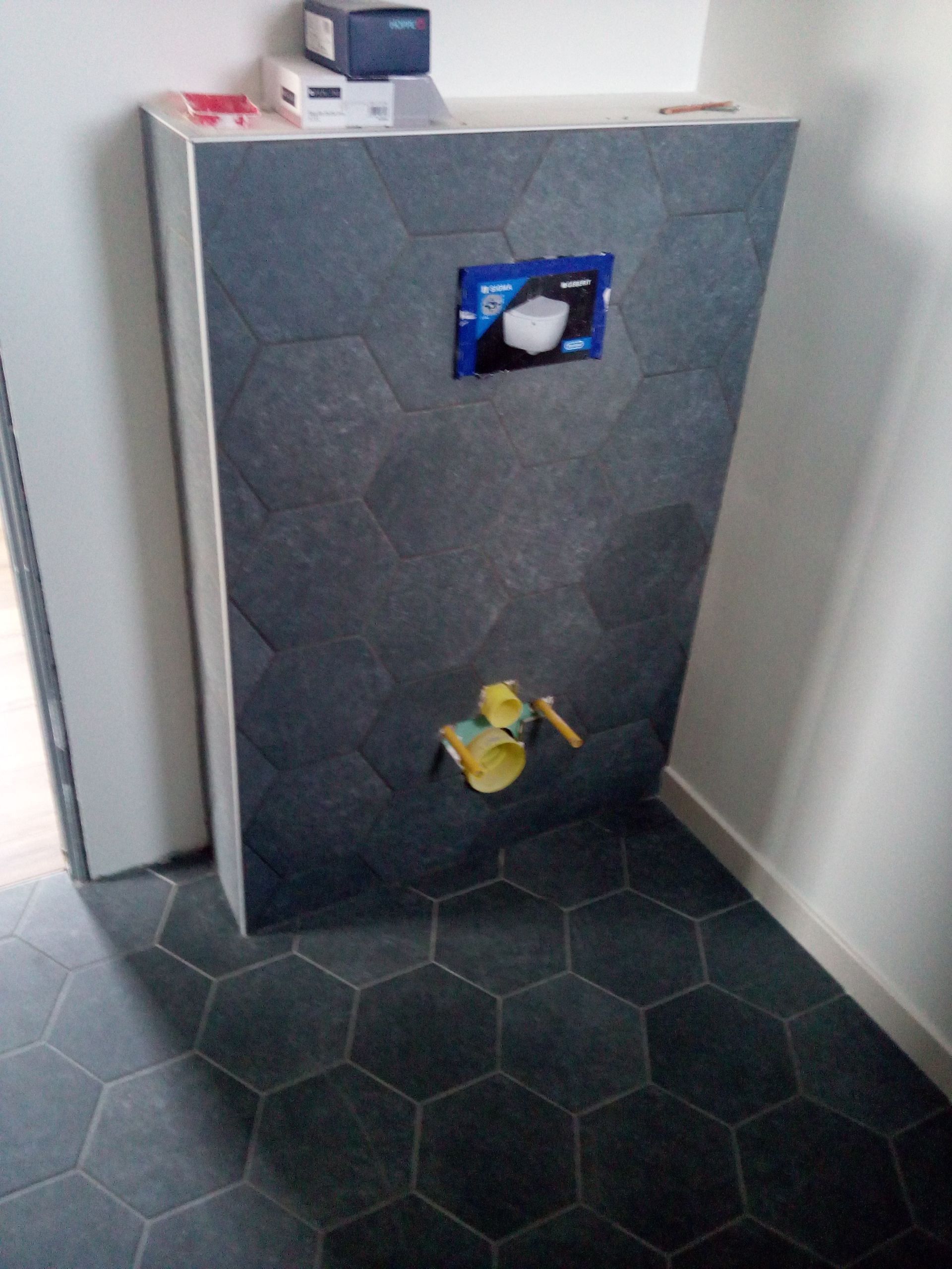 Installation de toilettes suspendues avec carrelage hexagonal foncé au sol et à mi-hauteur, comprenant un mécanisme de chasse d'eau bleu.