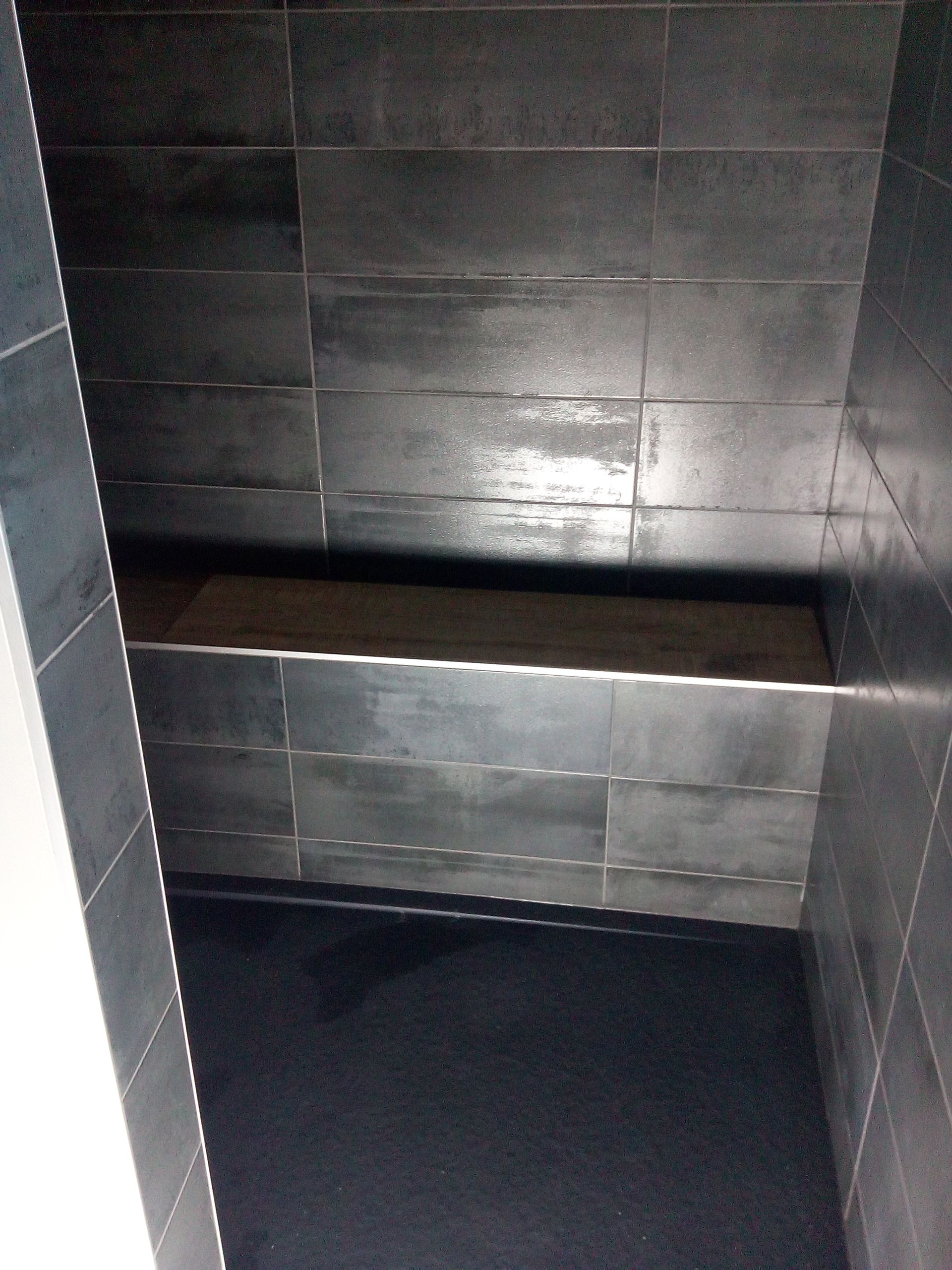 Une pièce faiblement éclairée, sans fenêtres, aux murs et au sol carrelés de gris foncé, avec une banquette carrelée intégrée.