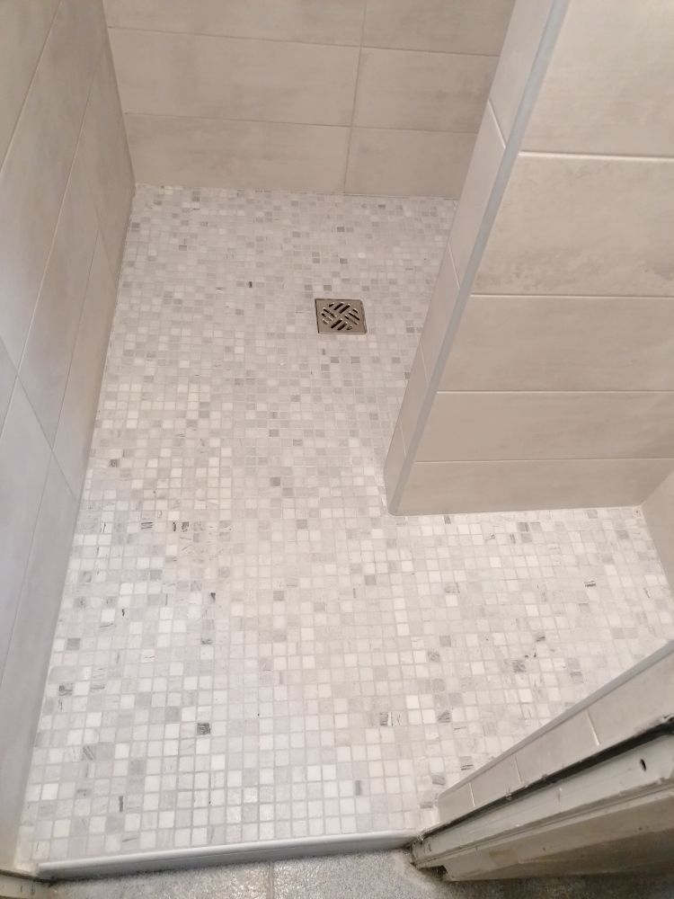 Une cabine de douche avec un sol en mosaïque de couleur claire, une bonde centrale et de grands carreaux muraux rectangulaires.