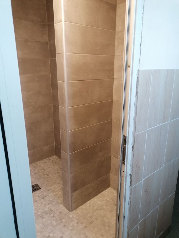 Une cabine de douche étroite carrelée, avec des carreaux muraux bruns et des carreaux de sol clairs, vue à travers une porte.
