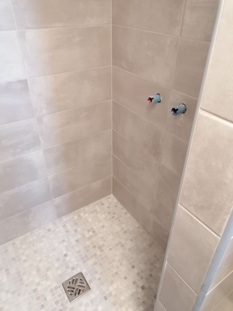 Une douche à l'italienne carrelée avec des murs gris clair, un sol en mosaïque, une bonde carrée et deux entrées de tuyaux non finies.