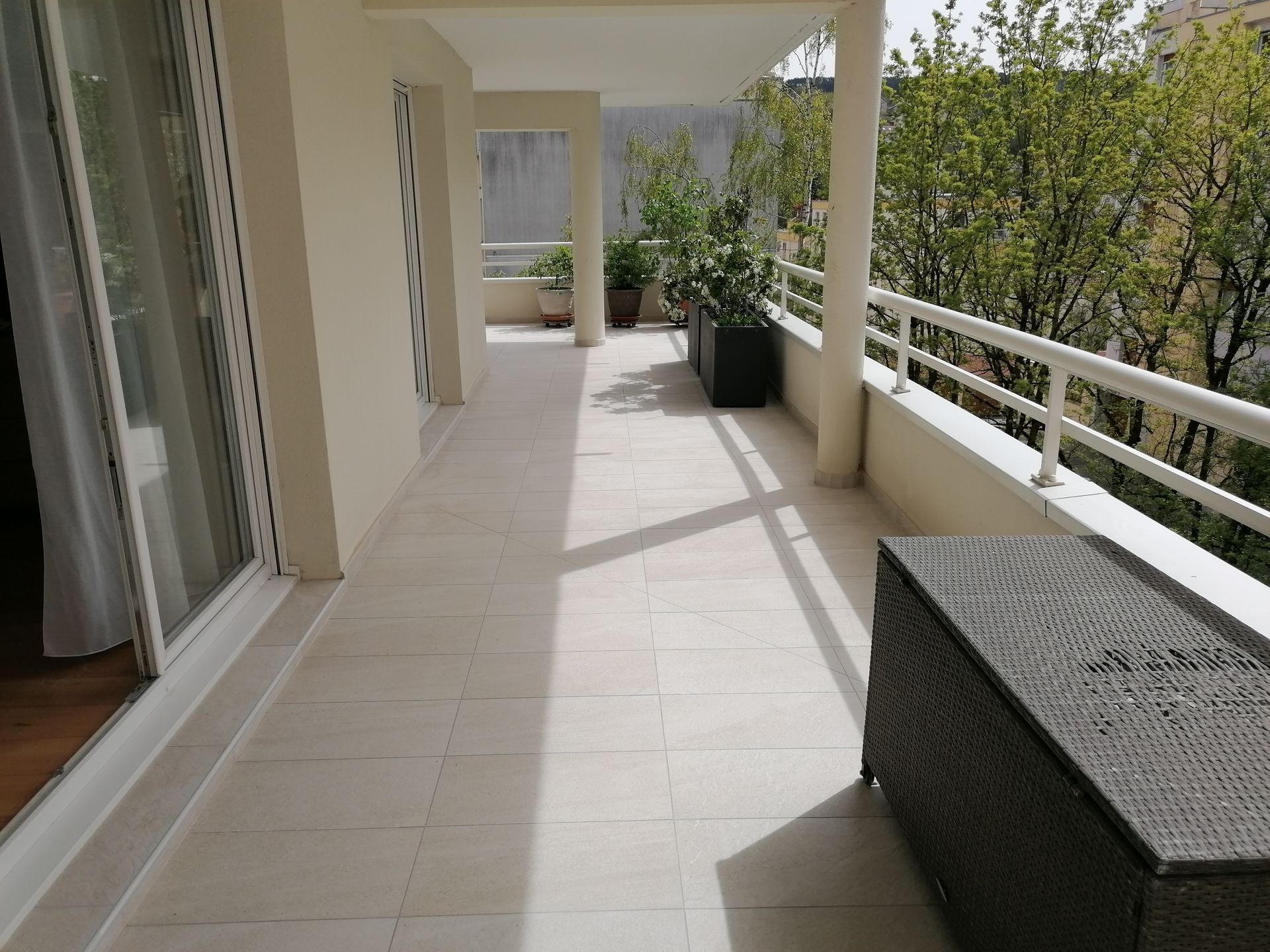 Un balcon ensoleillé avec un sol carrelé blanc, un coffre de rangement sombre au premier plan, des plantes en pot et une rambarde blanche.