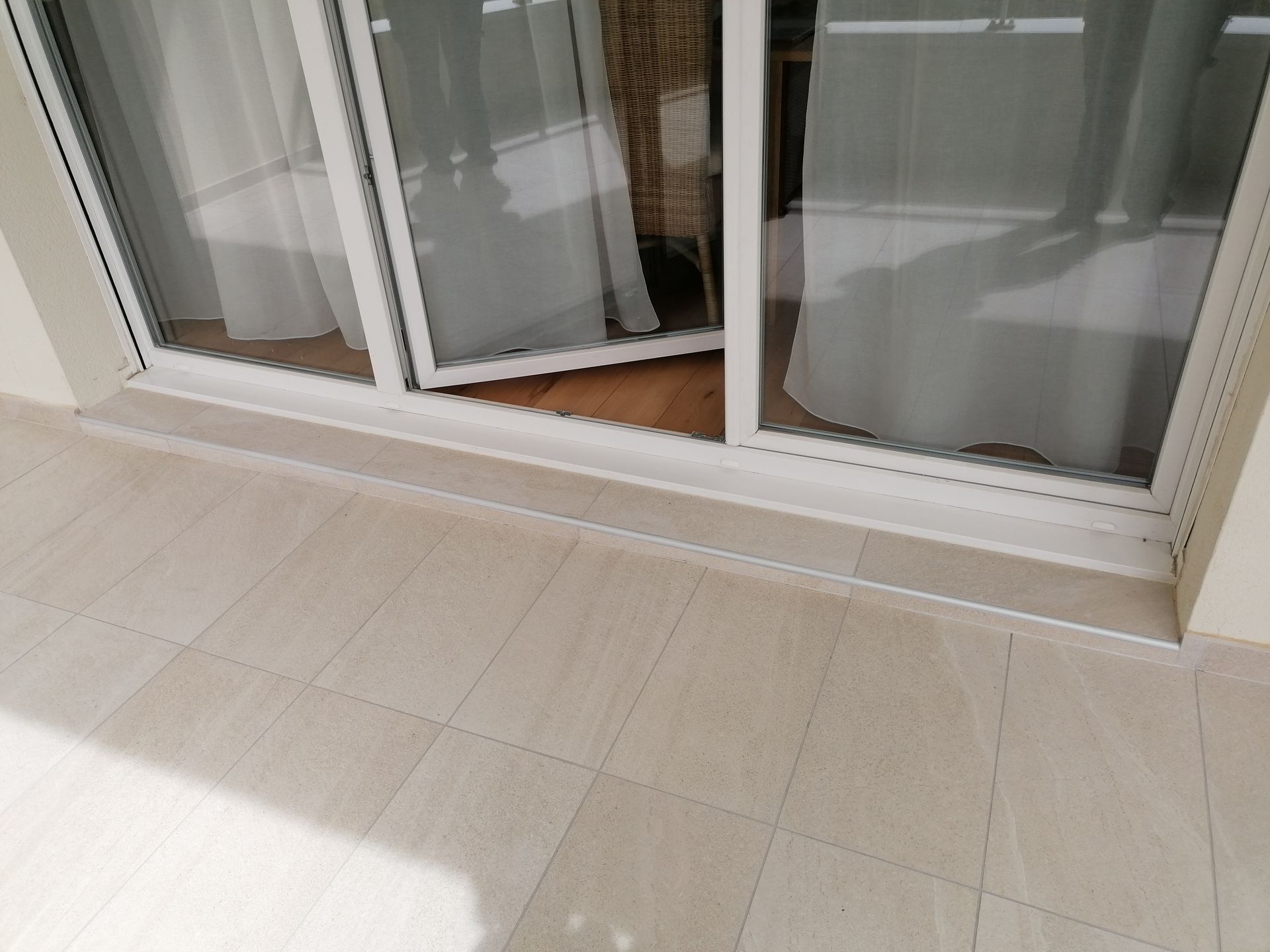 Une porte coulissante en verre s'ouvre sur un balcon carrelé de beige, laissant apparaître des rideaux blancs à travers les panneaux de verre.