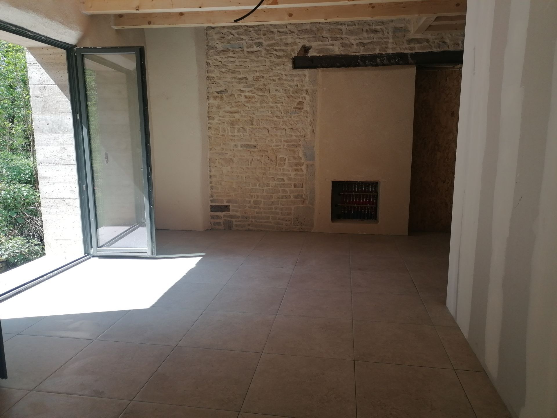 Intérieur d'une pièce avec murs en pierre, cheminée, grandes portes vitrées et sol carrelé clair.