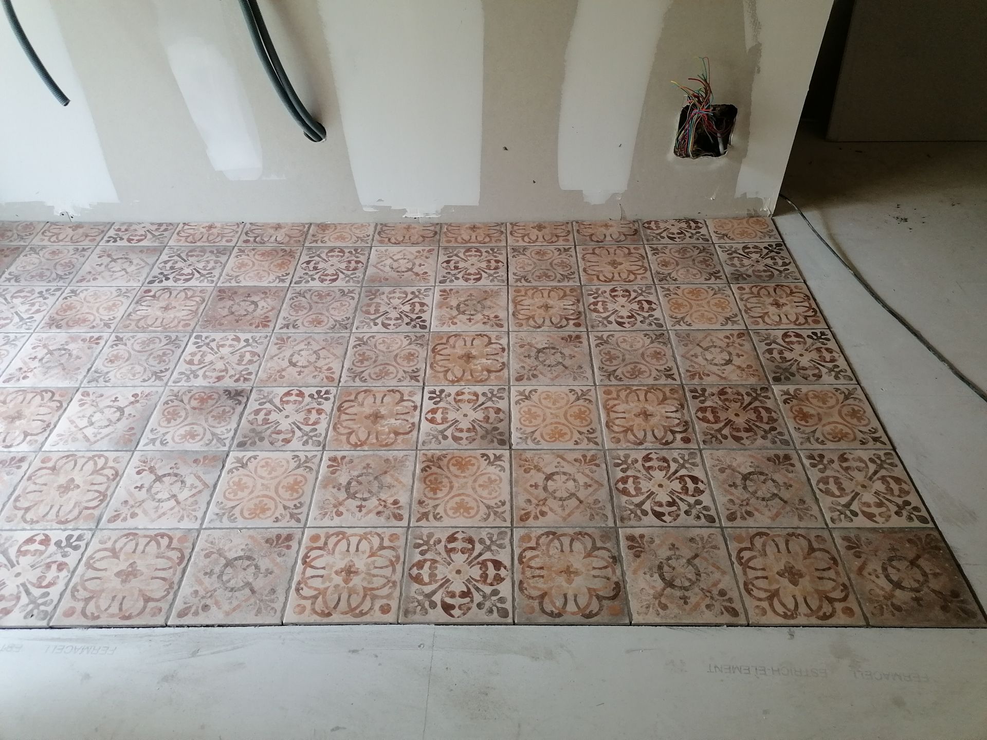 Carreaux carrés décoratifs à motif floral dans des tons de brun et de beige, posés sur le sol d'une pièce non finie.