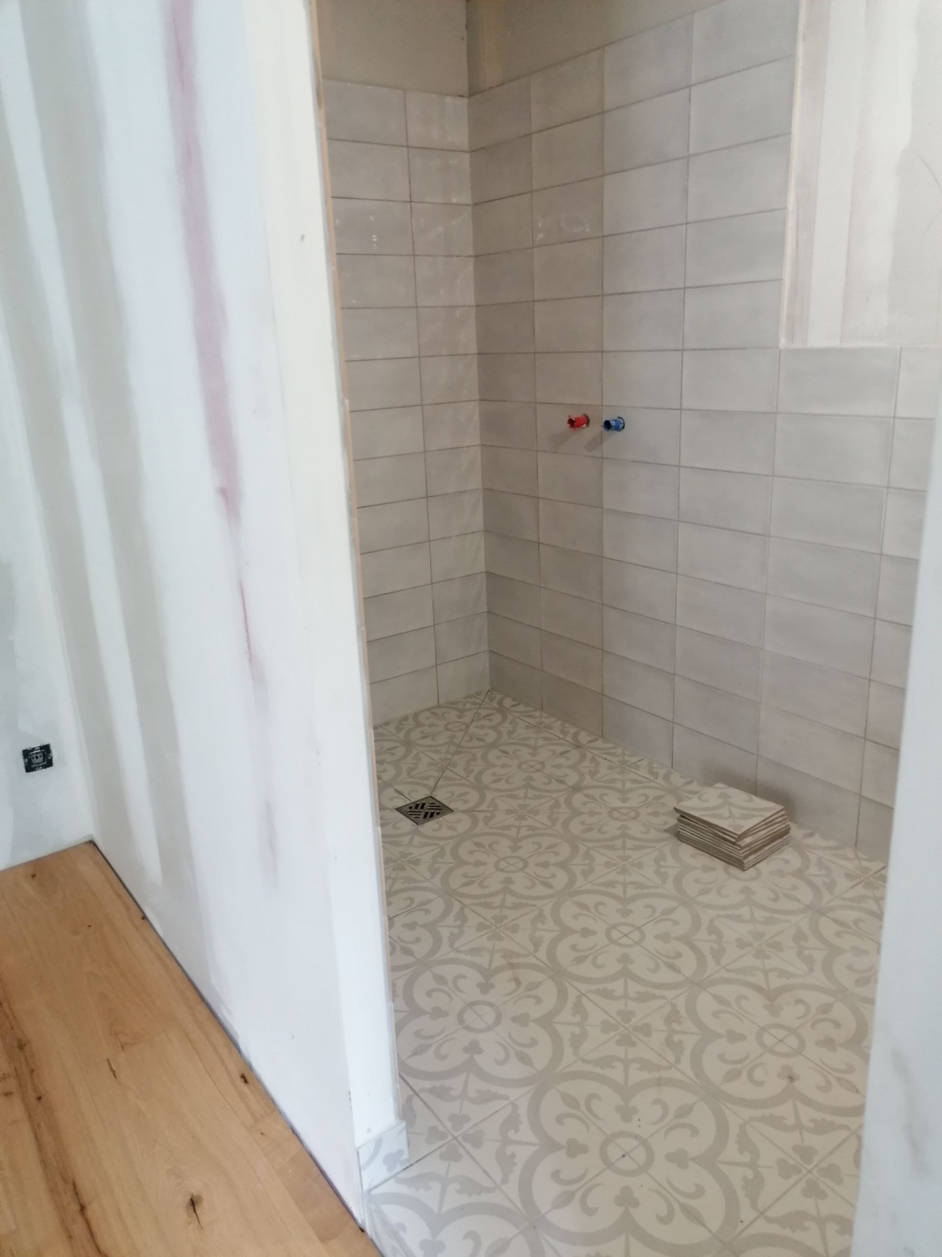 Une douche à l'italienne en construction, avec des carreaux muraux rectangulaires gris clair et des carreaux de sol à motifs.