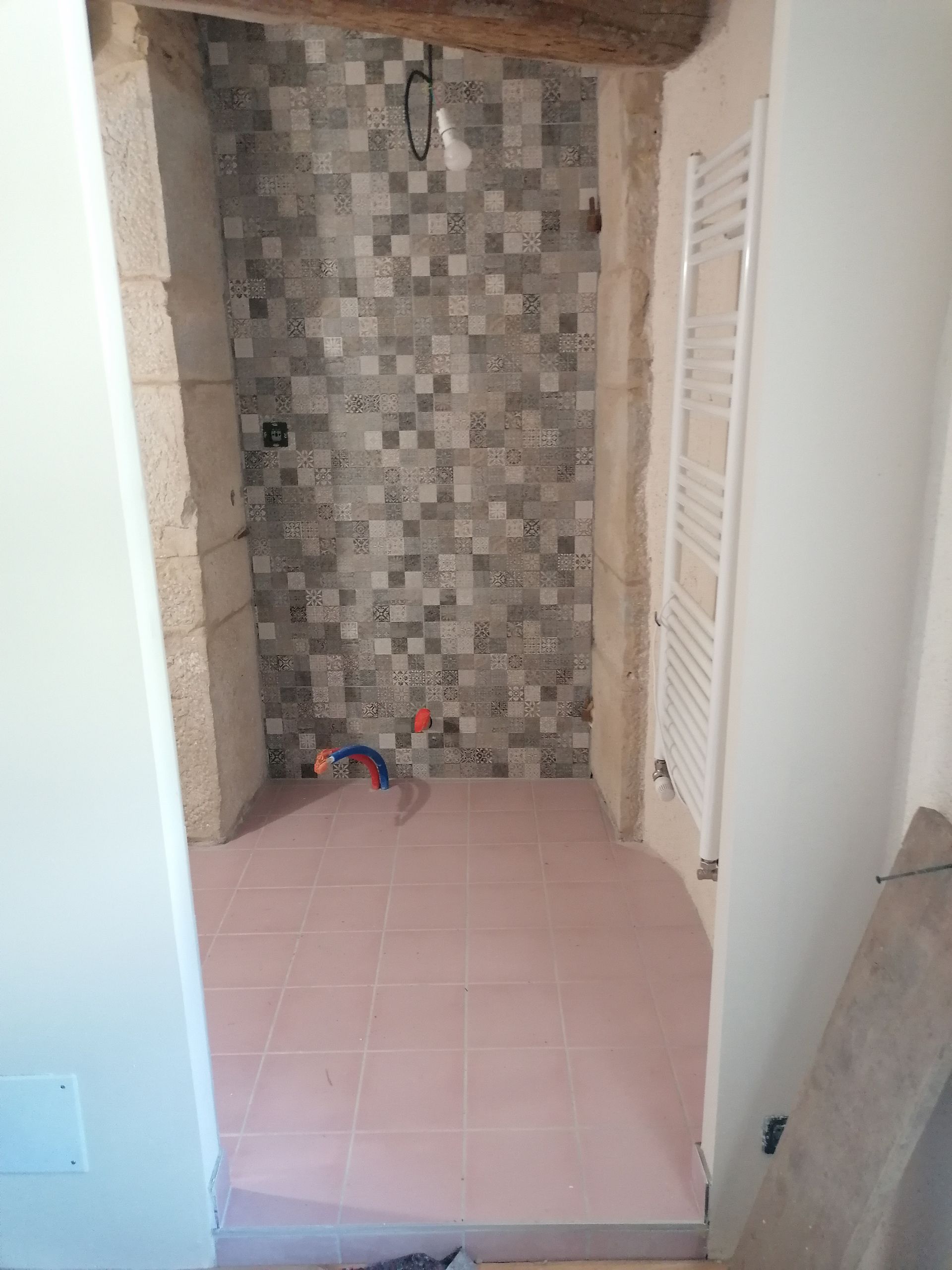Une douche partiellement rénovée avec des carreaux de mosaïque gris au fond, des carreaux de sol roses, une tuyauterie apparente et un sèche-serviettes blanc.