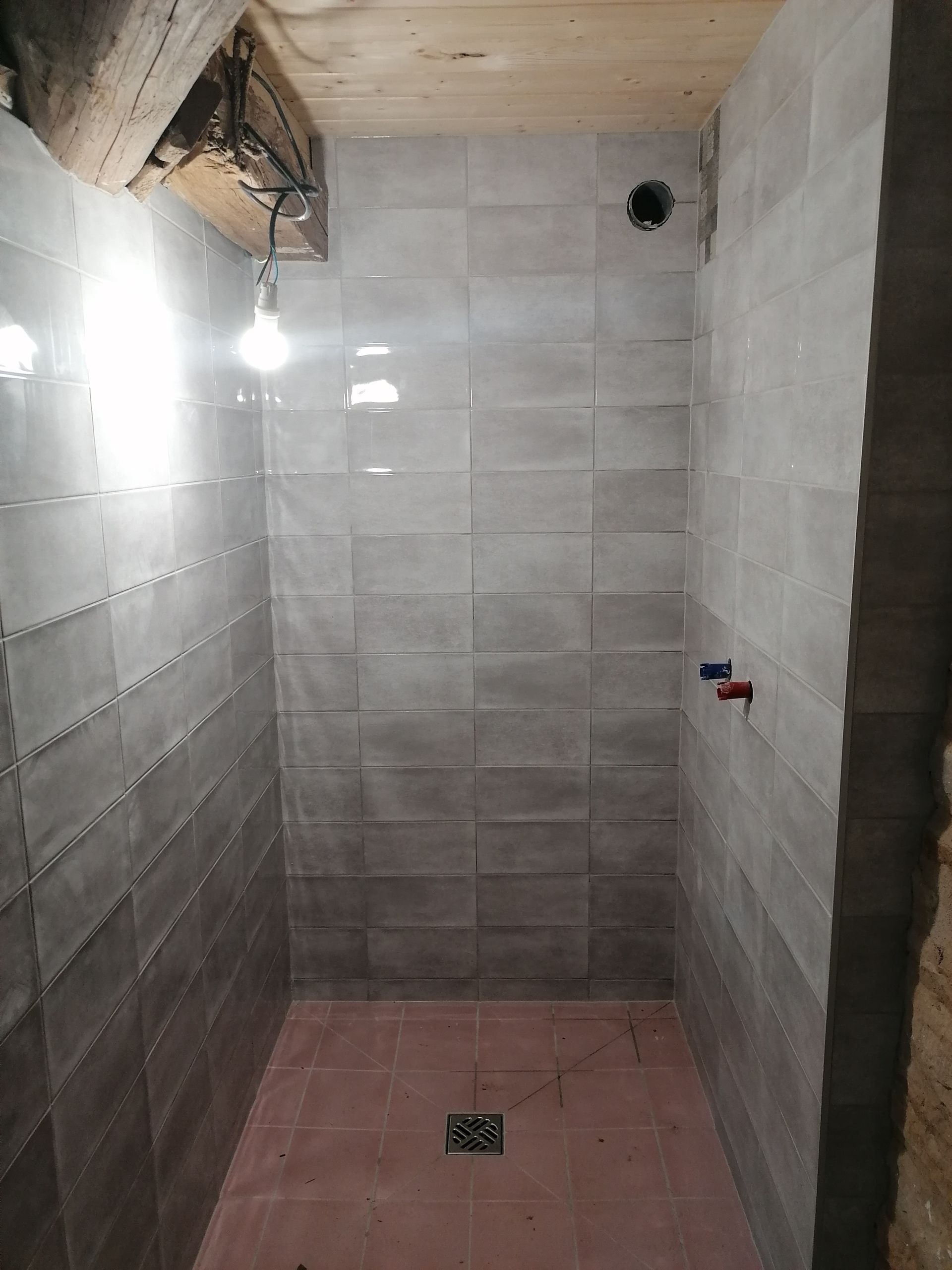 Une cabine de douche avec des carreaux muraux rectangulaires gris, un sol carrelé rose clair avec une bonde et un plafond à poutres apparentes en bois.