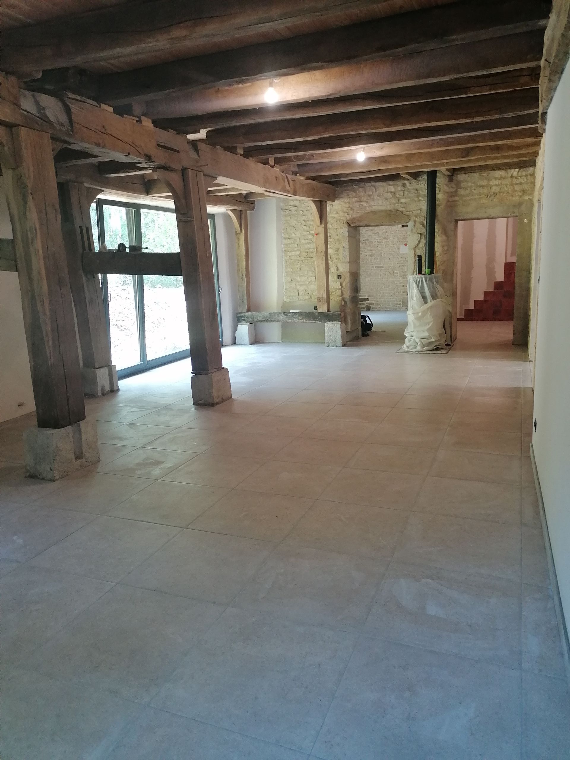 Une pièce ouverte avec des poutres en bois apparentes, des murs en pierre et un sol carrelé clair, donnant sur une porte avec un escalier.