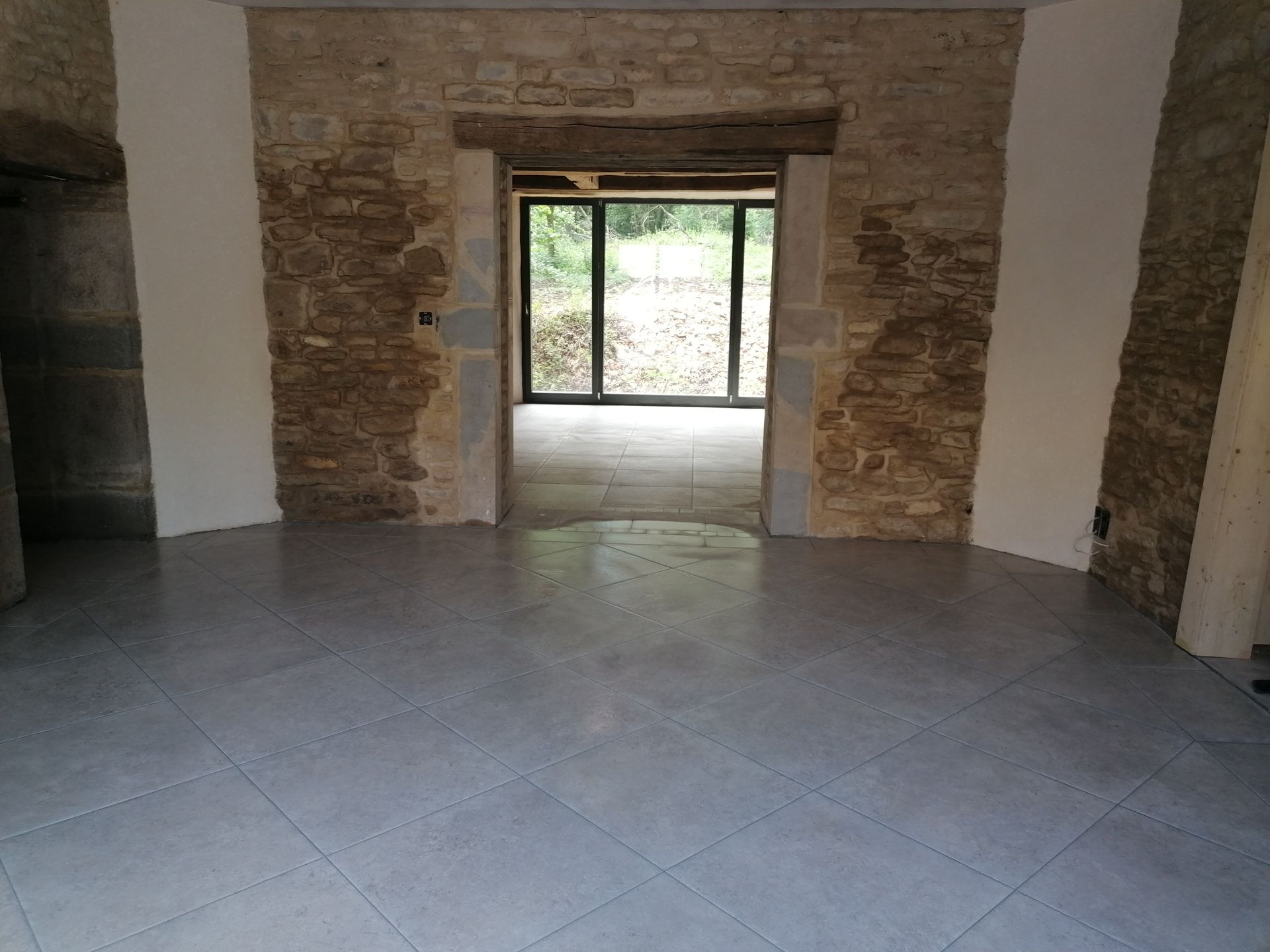 Une pièce avec un sol carrelé gris clair, des murs en pierre, des piliers blancs et une grande porte donnant sur un jardin.