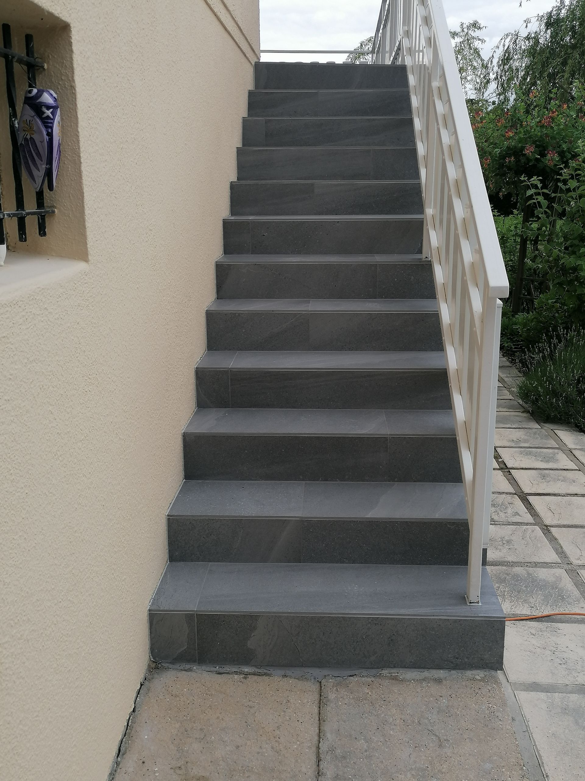 Un escalier extérieur droit en pierre grise menant à un mur beige, à côté d'une rambarde décorative en métal blanc.