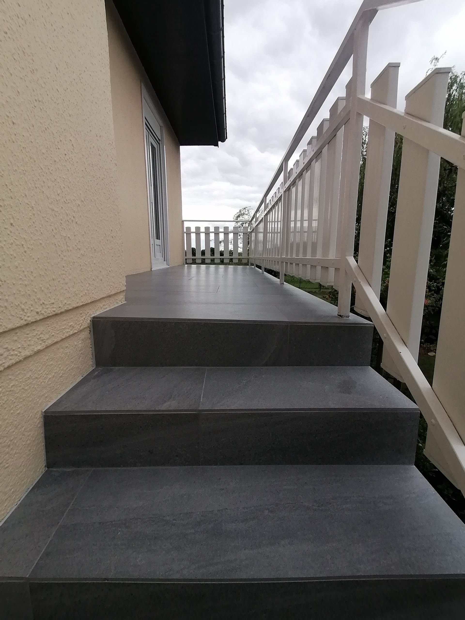 Un escalier extérieur carrelé de gris mène à une porte, avec une rambarde en bois blanc sur le côté.