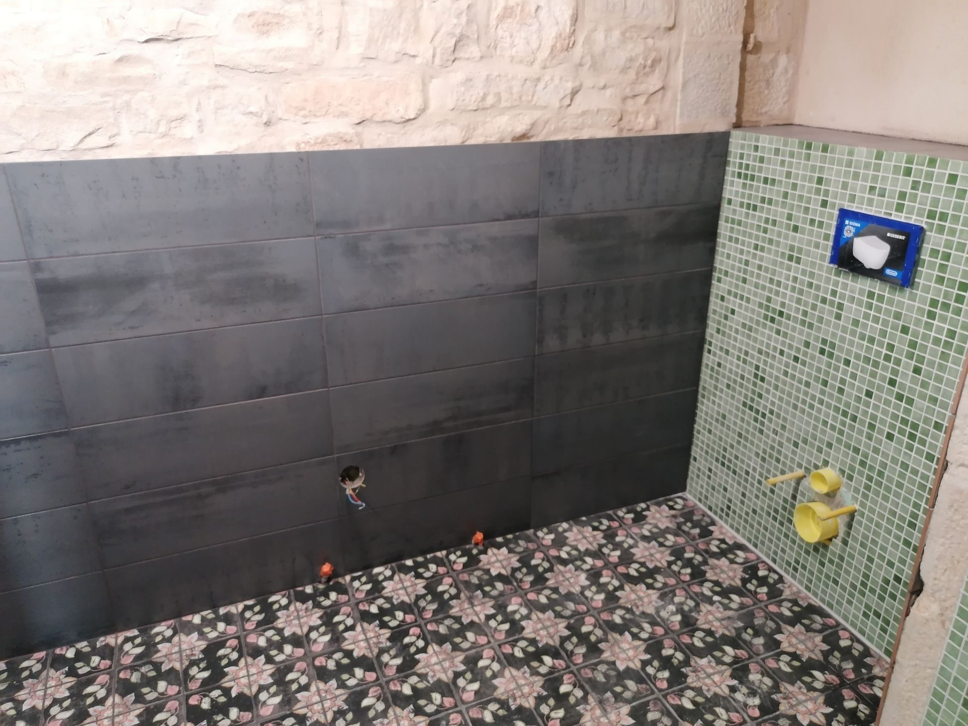 Une salle de bains en construction avec des carreaux horizontaux noirs, un mur en mosaïque verte avec une plaque de commande et un sol à motifs.