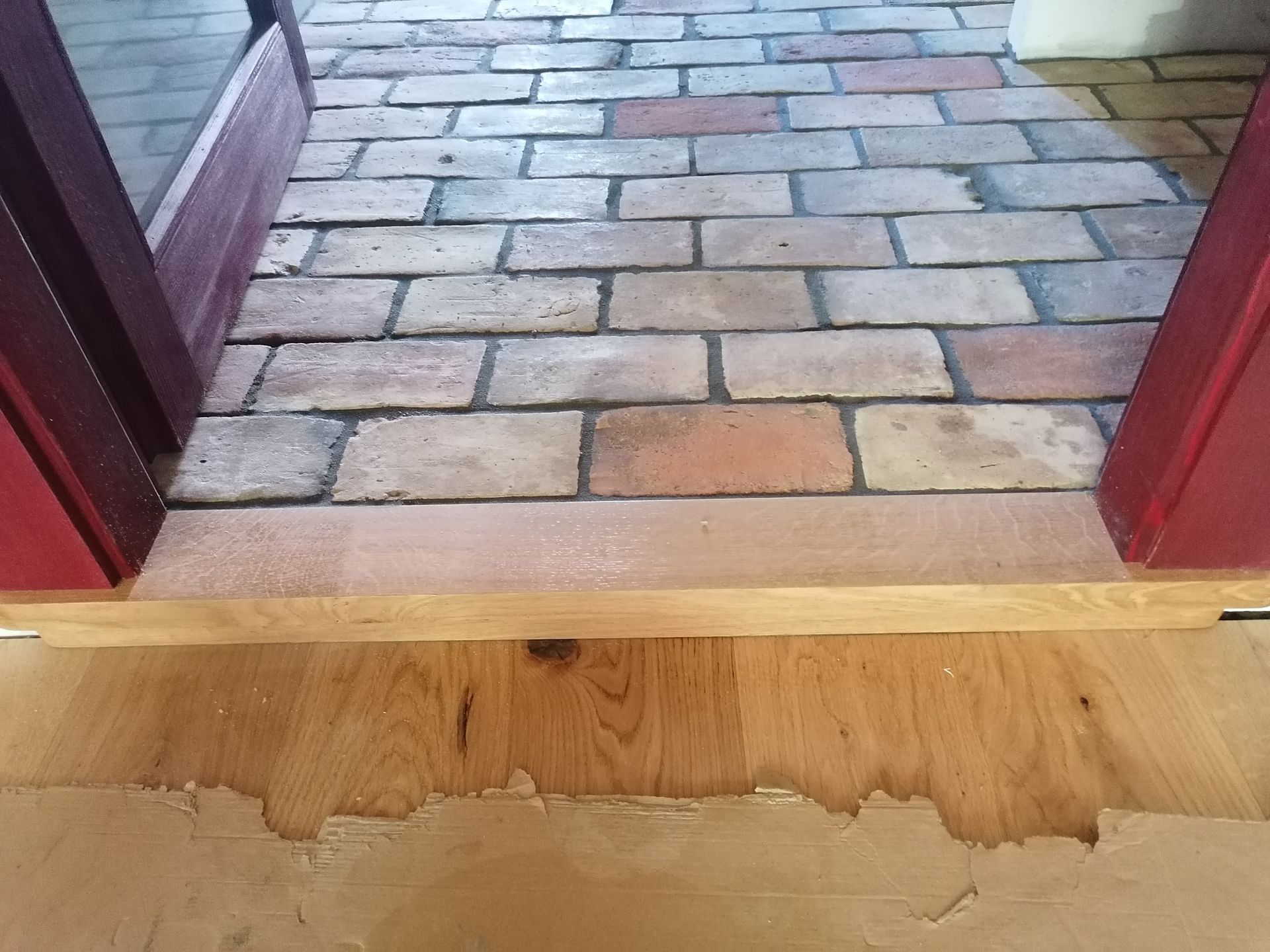 Un seuil en bois fait la transition entre un sol en briques rouges et un parquet clair recouvert d'une partie en papier.
