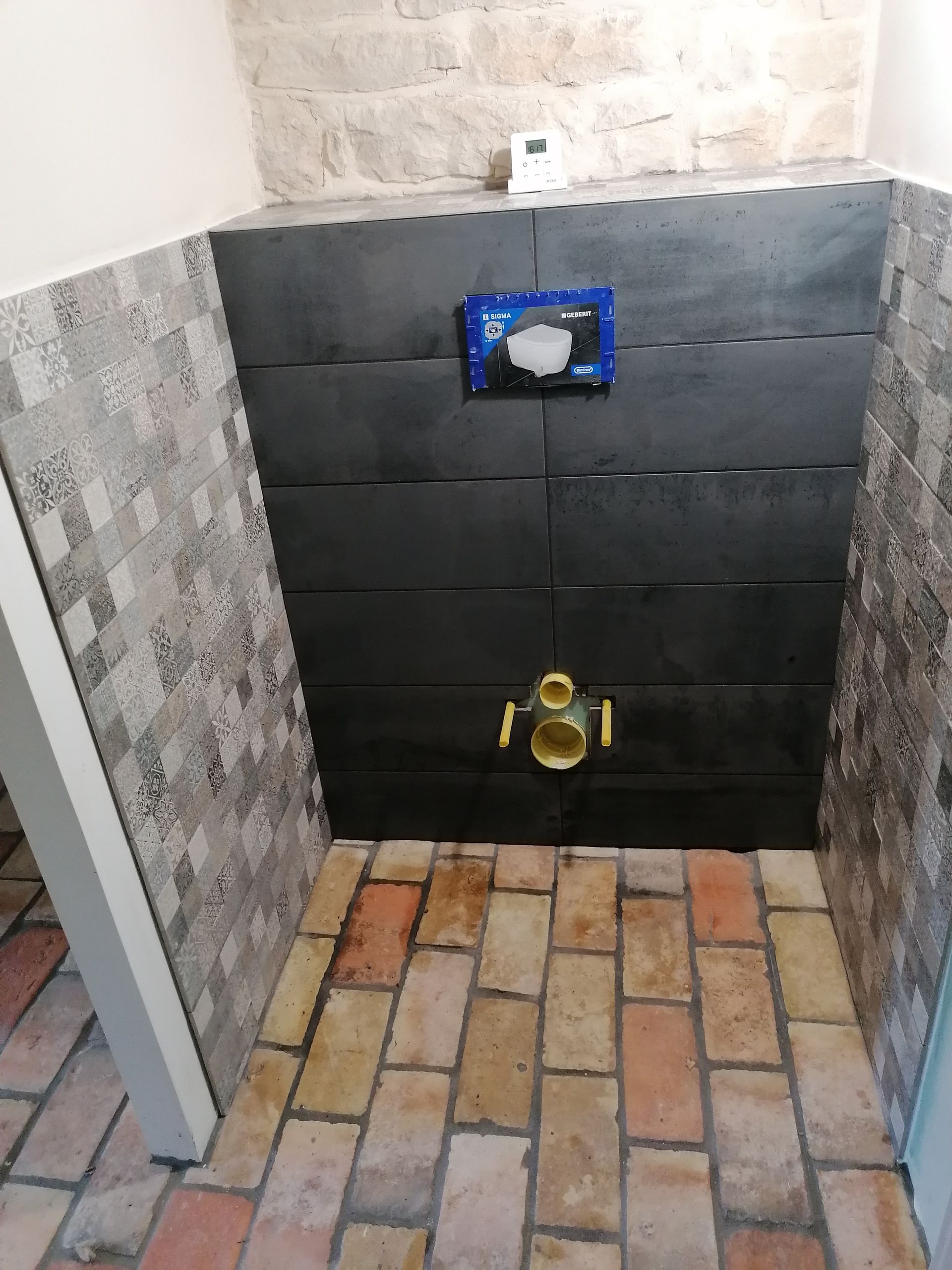 Un bâti-support mural pour WC avec tuyauterie apparente et plaque de commande, le tout sur un sol en carrelage foncé et en briques.