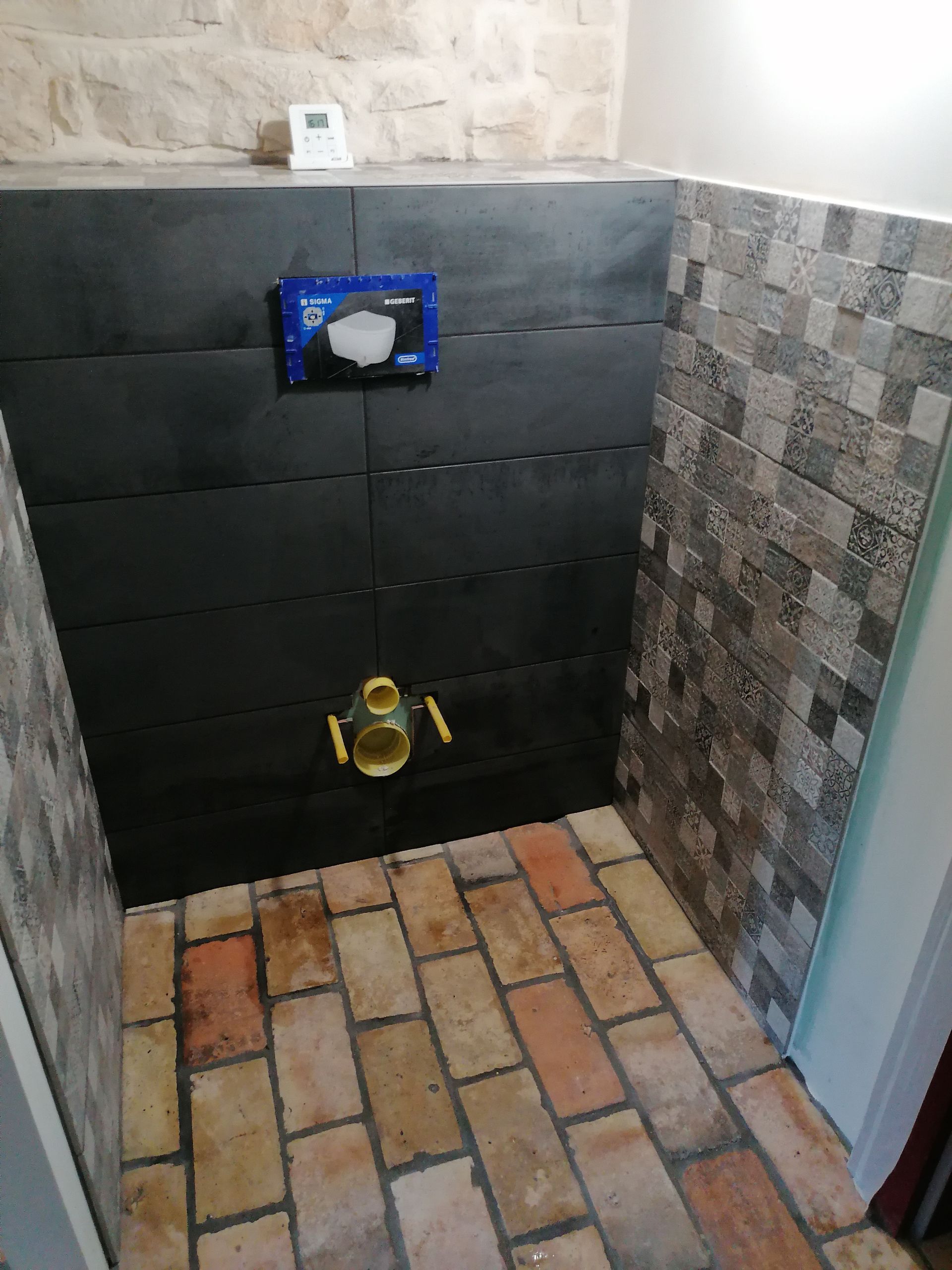 Un mur de salle de bains en cours de réalisation, avec des carreaux horizontaux foncés, une chasse d'eau encastrée et un sol à motif de briques.