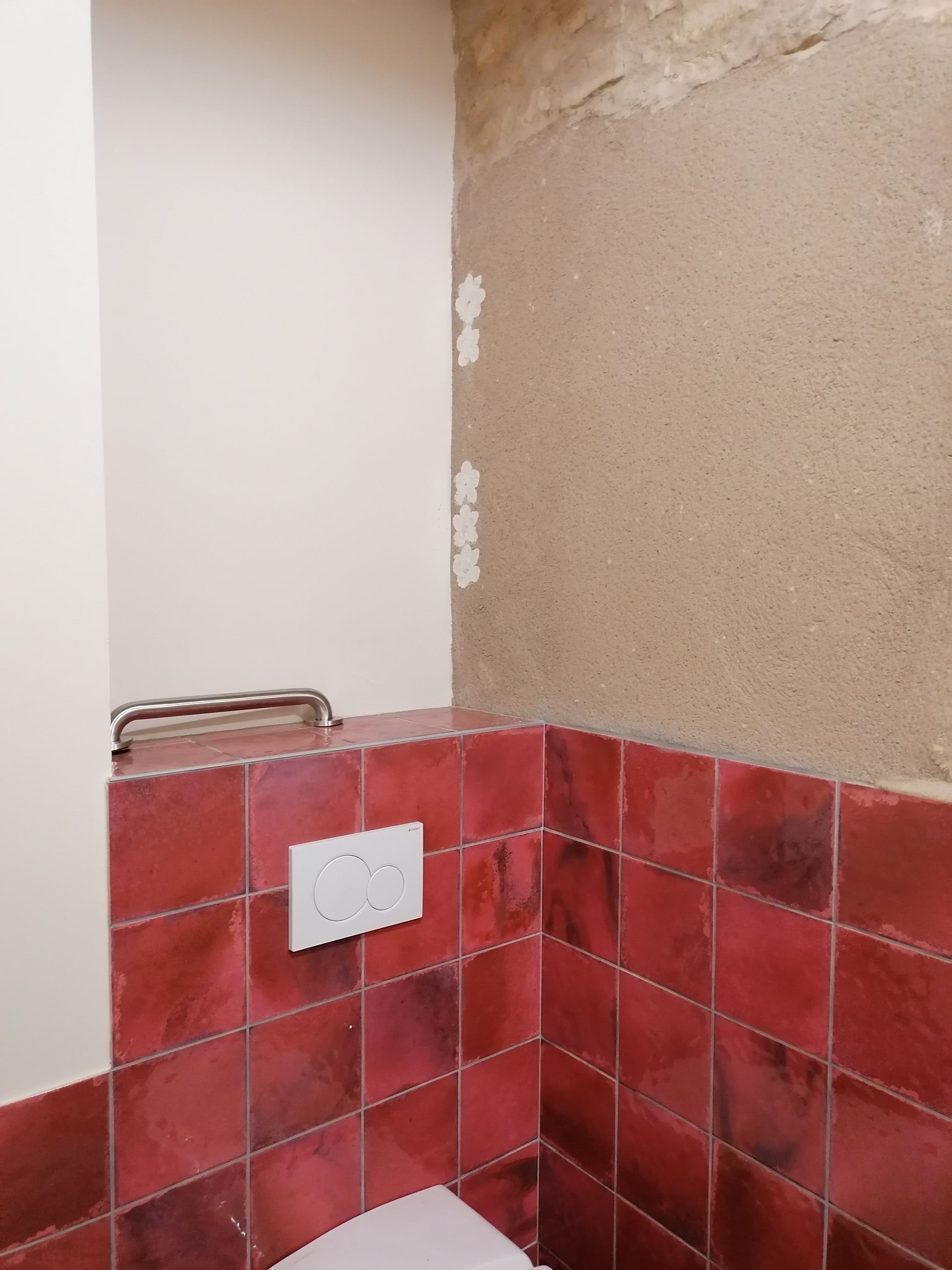 Vue d'angle d'une salle de bains montrant un mur carrelé de carreaux carrés rouges, une plaque de chasse d'eau blanche et des zones murales non finies.