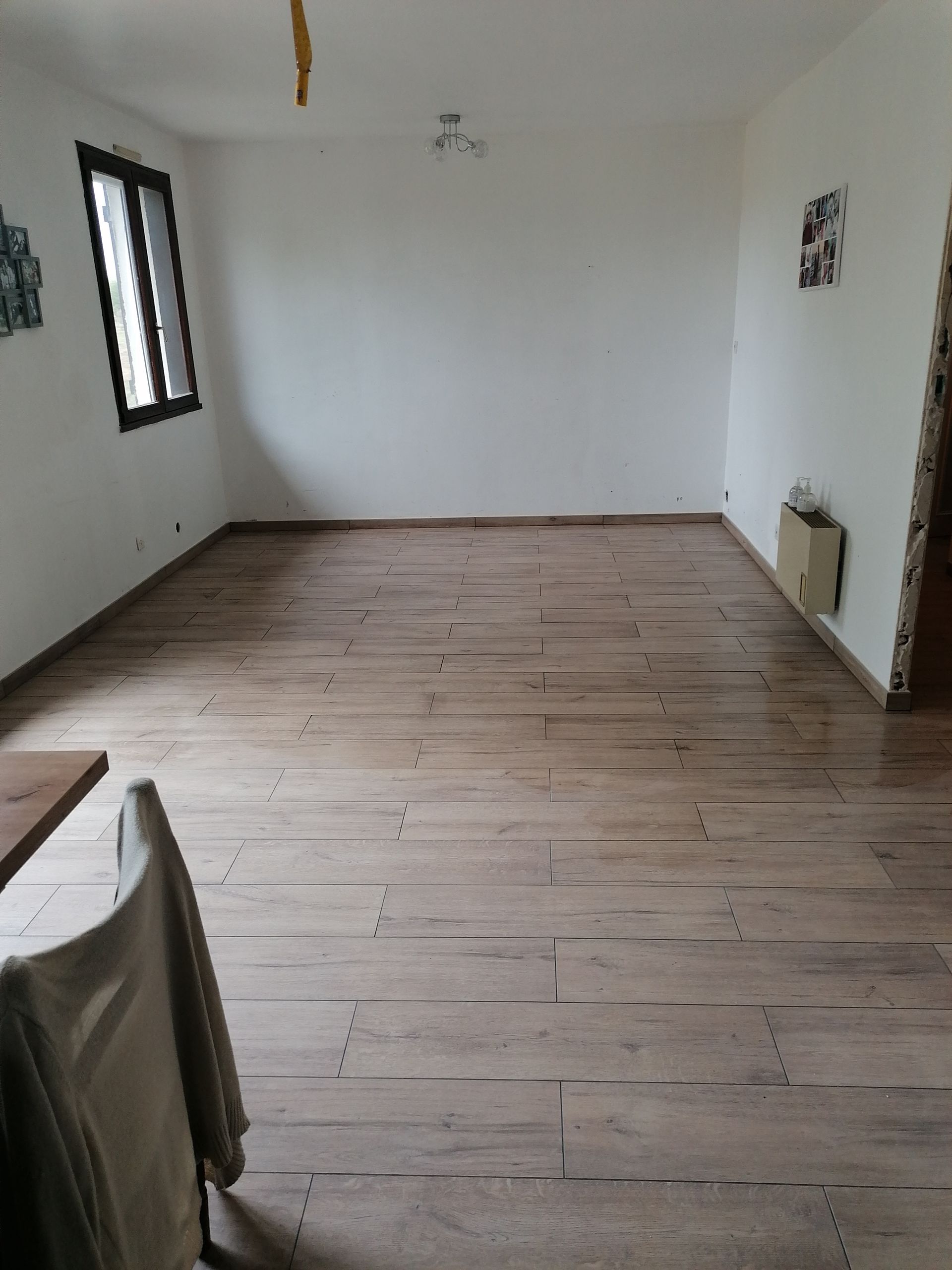 Une pièce vide avec un sol clair imitant le bois, des murs blancs, une fenêtre unique sur la gauche et un radiateur sur le mur de droite.