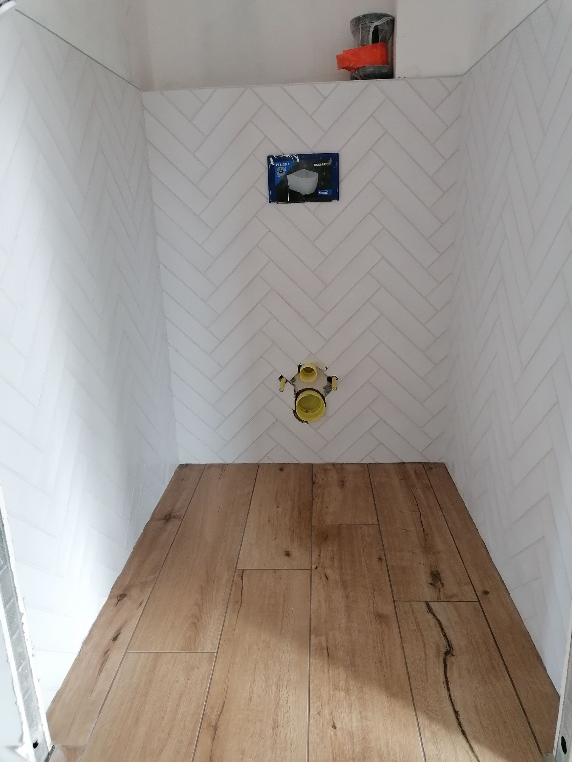 Une salle de bains en construction avec des carreaux muraux blancs à chevrons, des carreaux de sol imitation bois et une plomberie apparente.