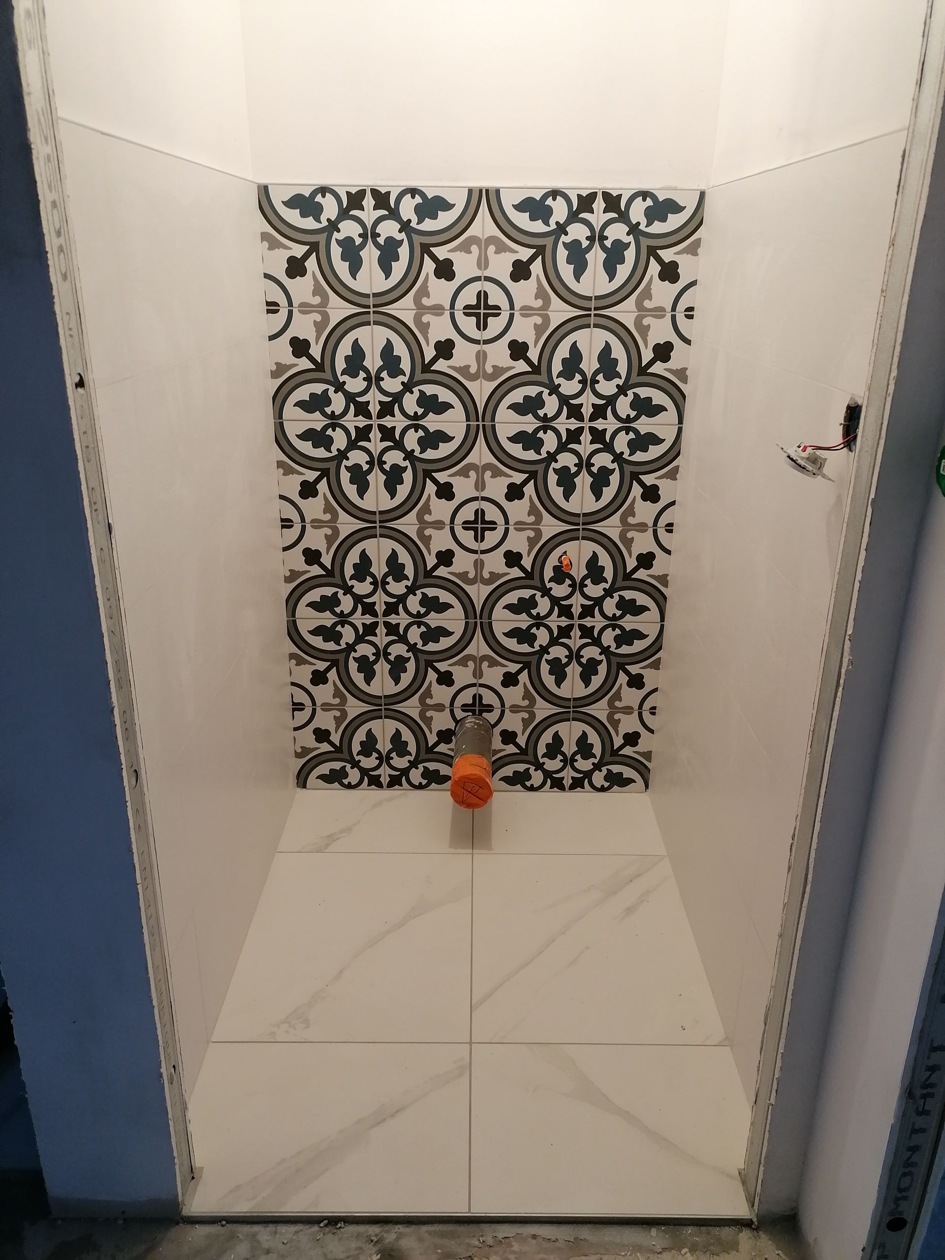 Une alcôve de douche carrelée avec un sol en carrelage imitation marbre blanc et un mur du fond orné de carreaux décoratifs à motifs floraux.