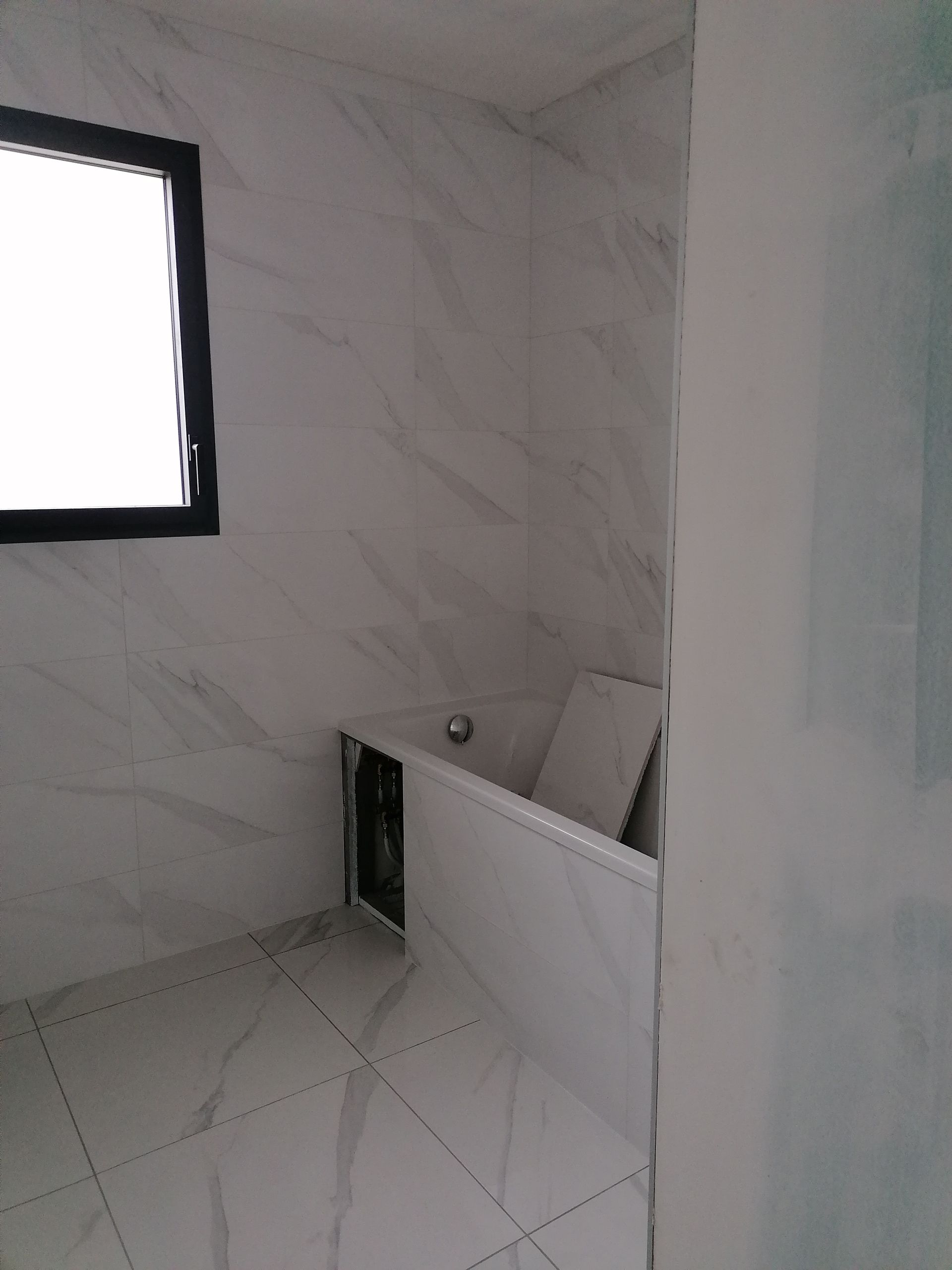 Une salle de bains moderne et lumineuse avec des carreaux muraux et de sol à motifs de marbre blanc, une fenêtre et une baignoire en construction.