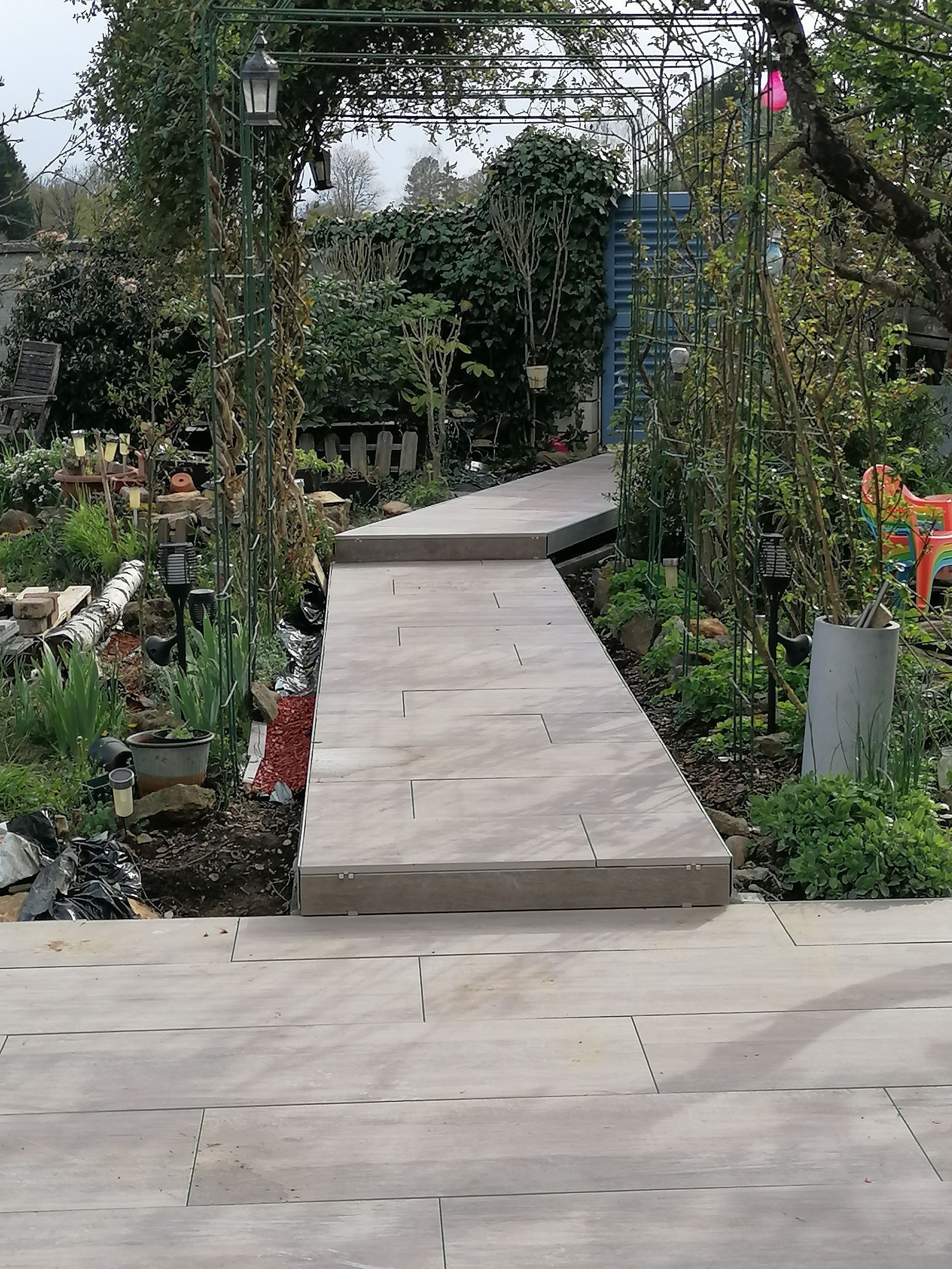 Allée de pierre traversant un jardin, menant à une arche métallique. Un feuillage vert borde le chemin.