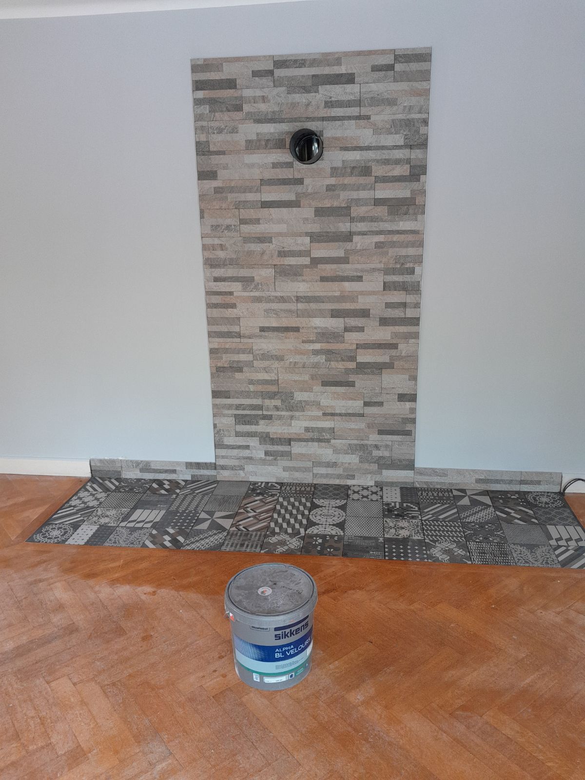 Un mur d'accent avec un carrelage vertical imitation pierre et une plinthe horizontale à motifs foncés, avec un seau de peinture sur un sol en bois.