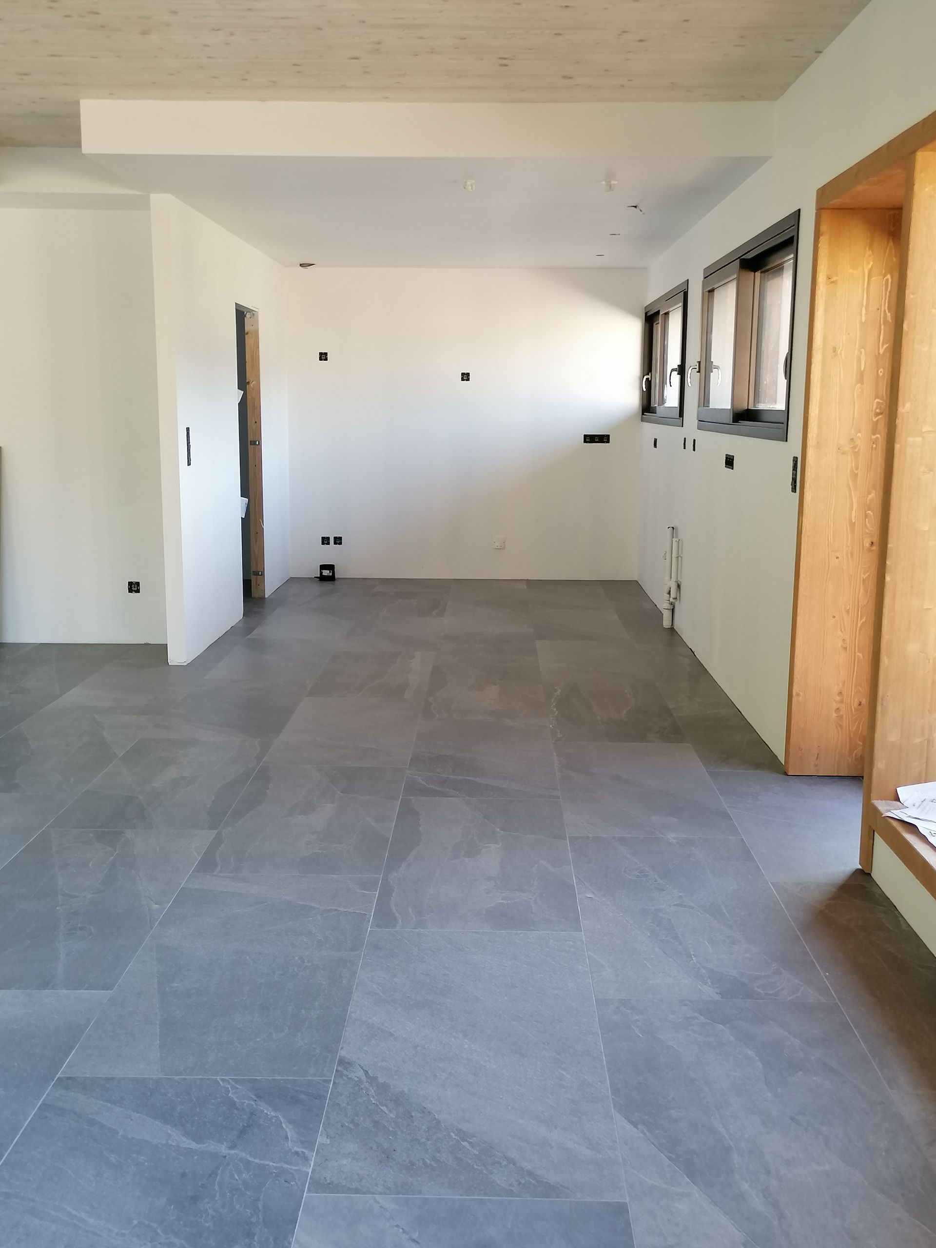 Une pièce vide avec un sol en pierre grise, des murs blancs et des ouvertures de fenêtres à cadre en bois.