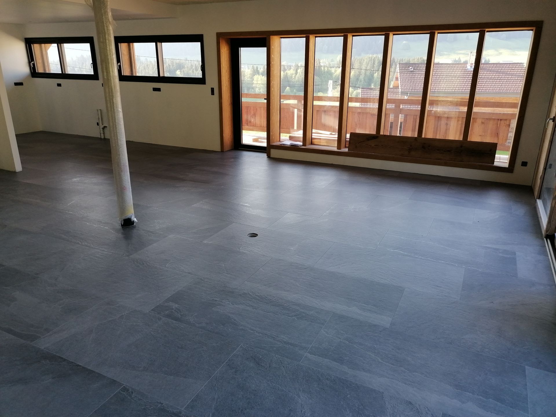 Une pièce lumineuse et vide, avec un sol carrelé gris, une simple colonne blanche et de grandes fenêtres donnant sur une terrasse.