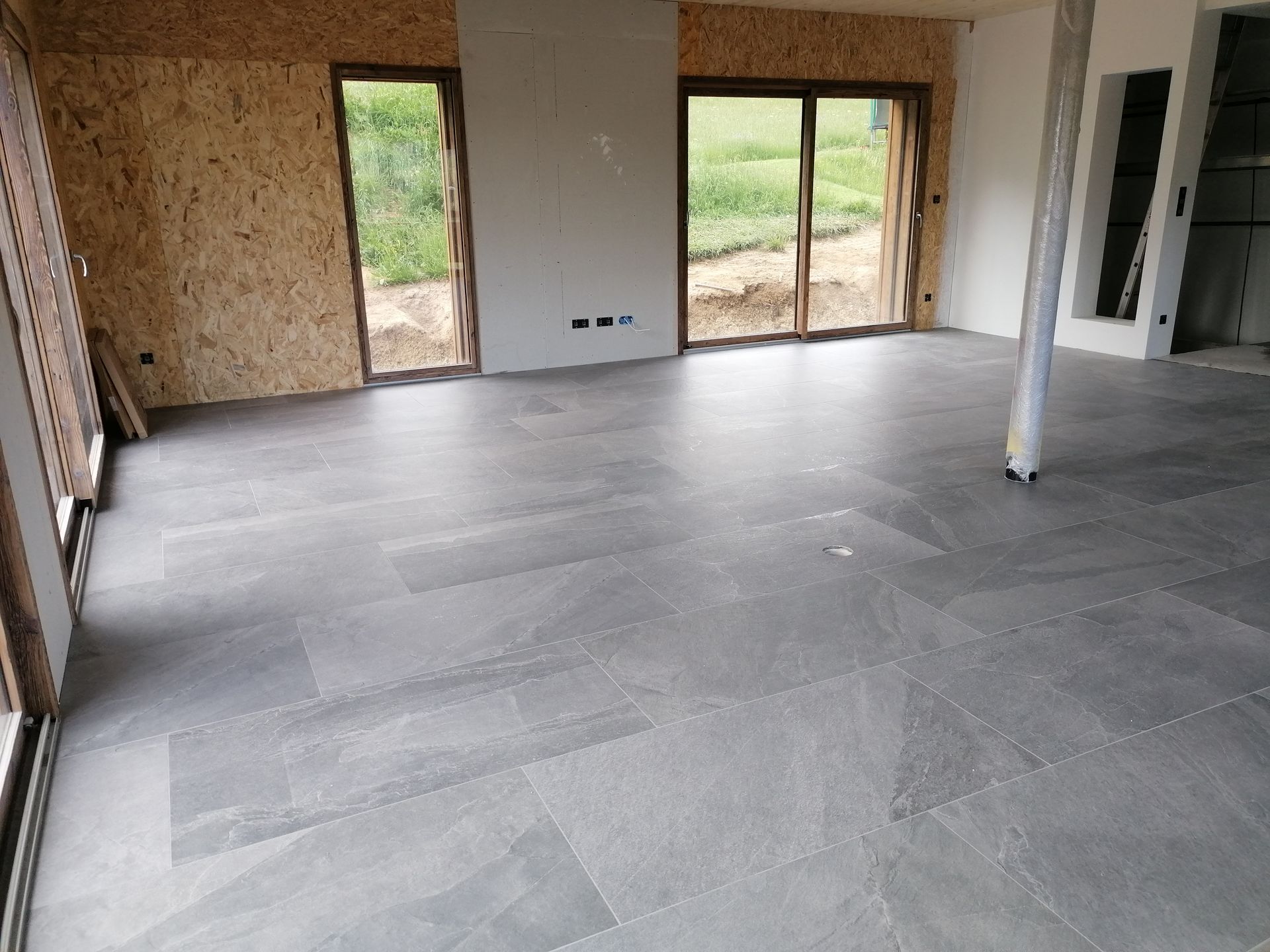 Pièce intérieure en construction avec sol carrelé gris, murs en bois clair et grandes fenêtres.