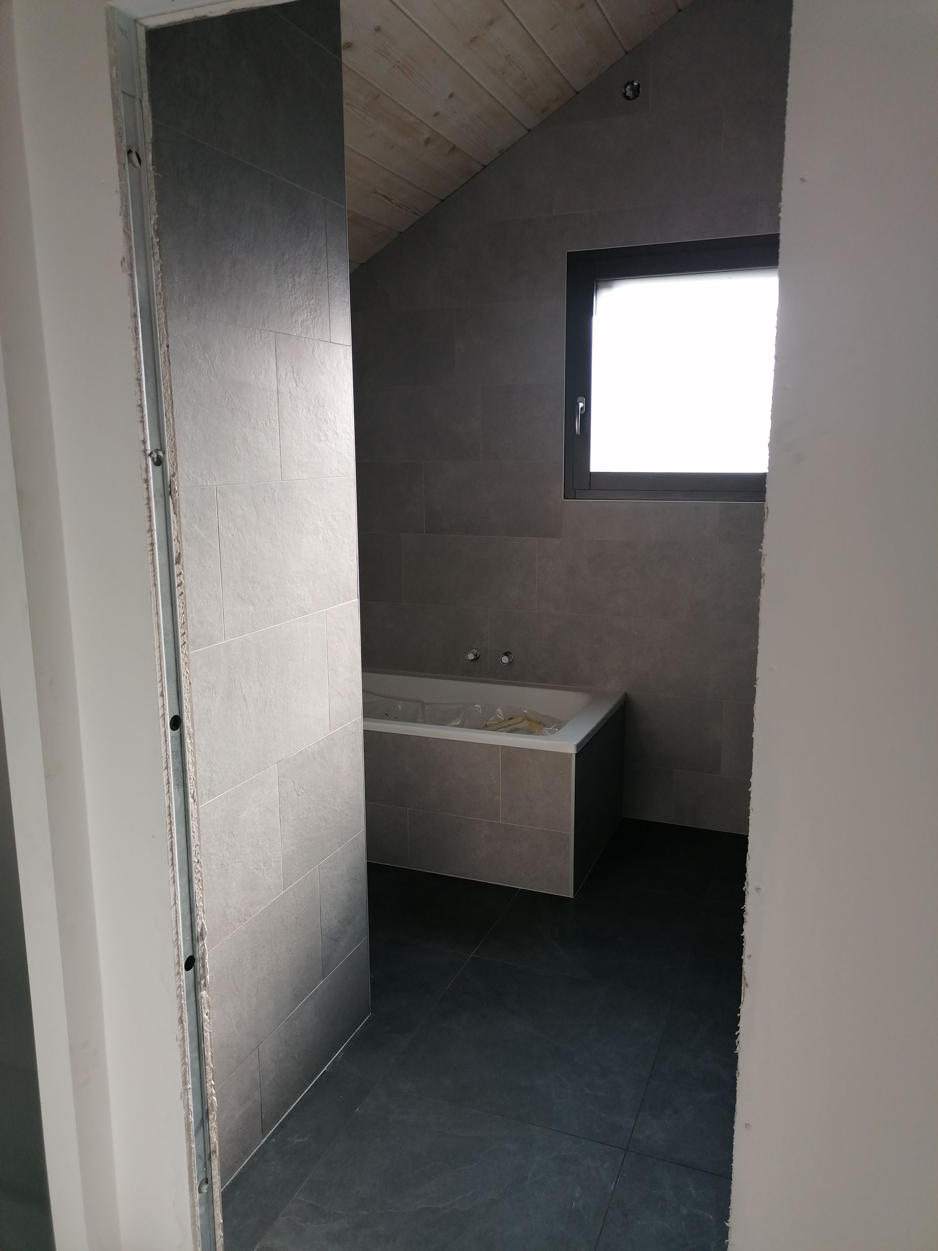 Vue à travers une porte donnant sur une salle de bains moderne avec des murs carrelés foncés, un sol en pierre grise et une baignoire encastrée.