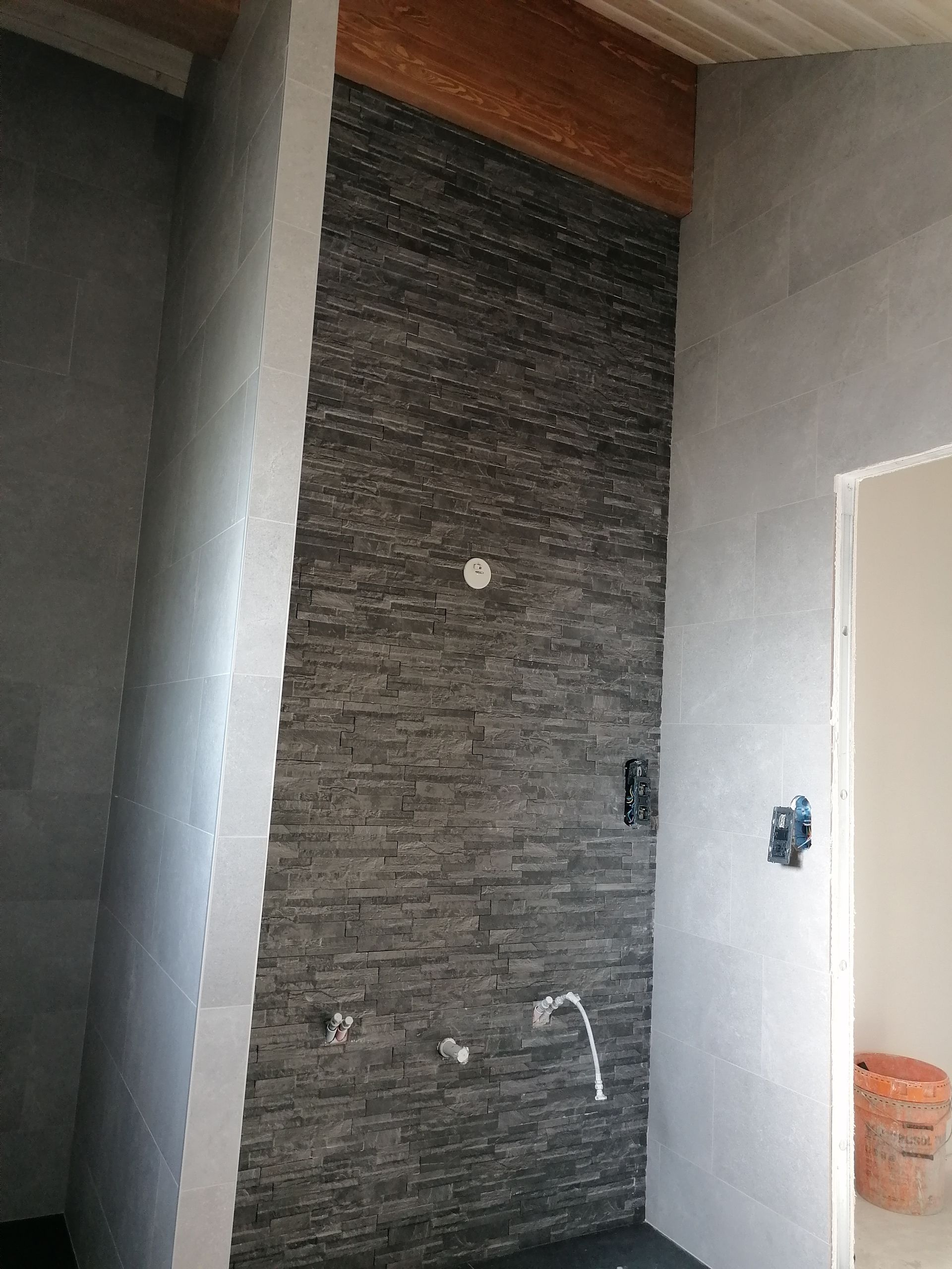 Une douche en construction avec un mur d'accent en pierre texturée gris foncé, des côtés carrelés gris clair et des tuyaux apparents.